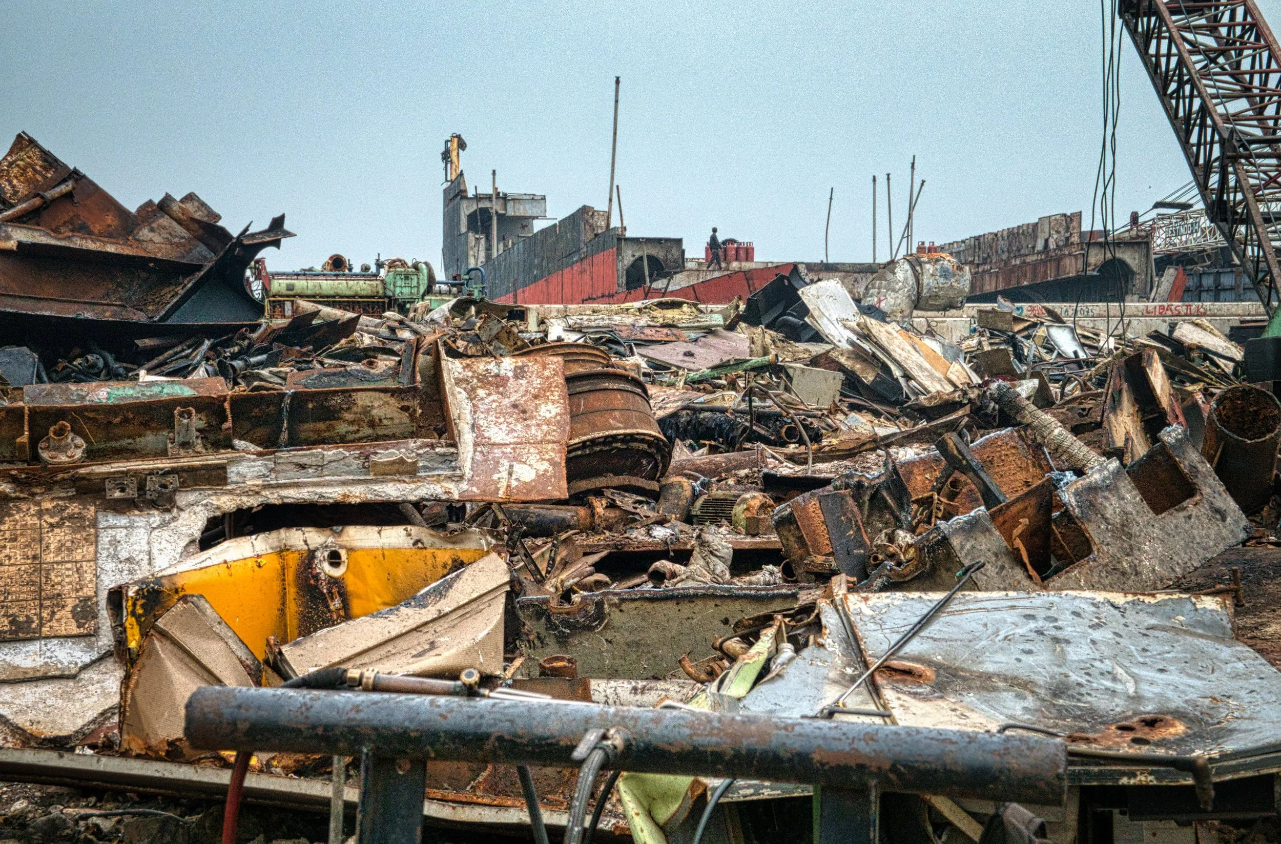 Large pile of rusted metal debris and scrap in a junkyard with a crane on the right side and a ship in the background.