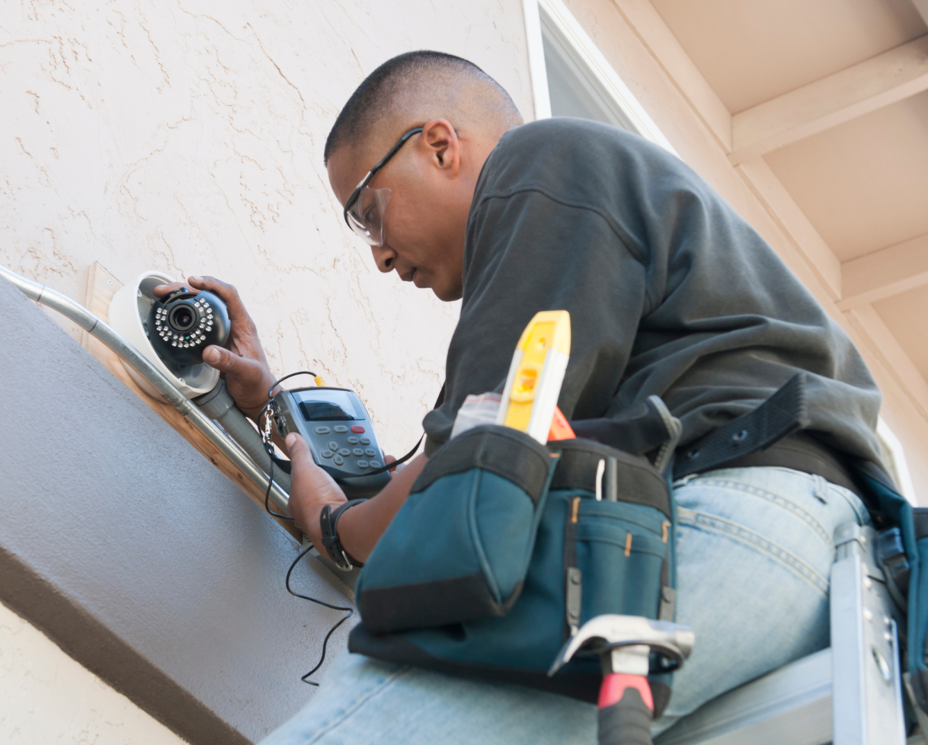 A technician installing a security camera on a house wall while working from a ladder, holding a special tool and a digital device.