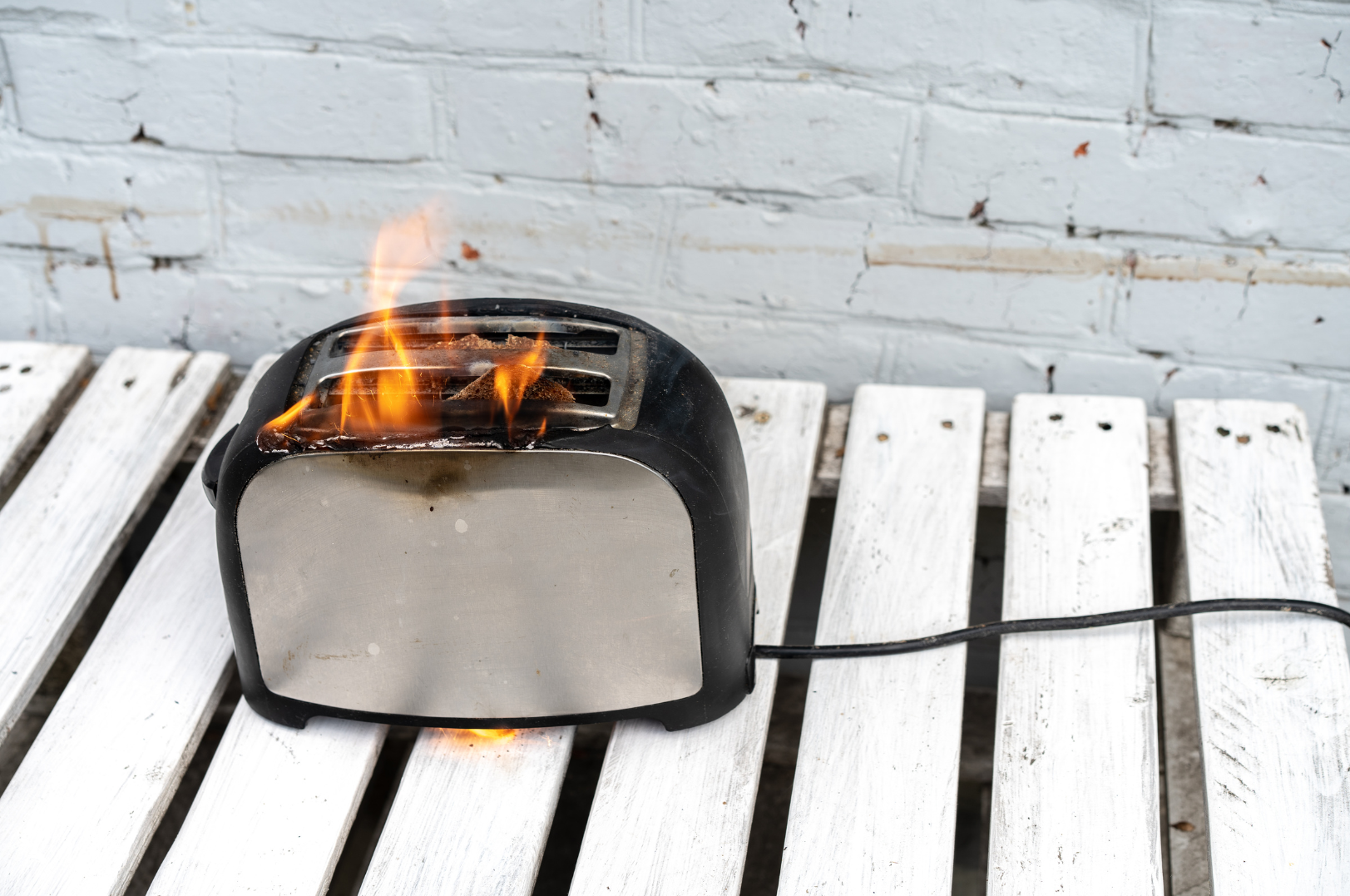 A black and gray toaster with a piece of bread on fire inside, placed on a white wooden surface against a white brick wall.
