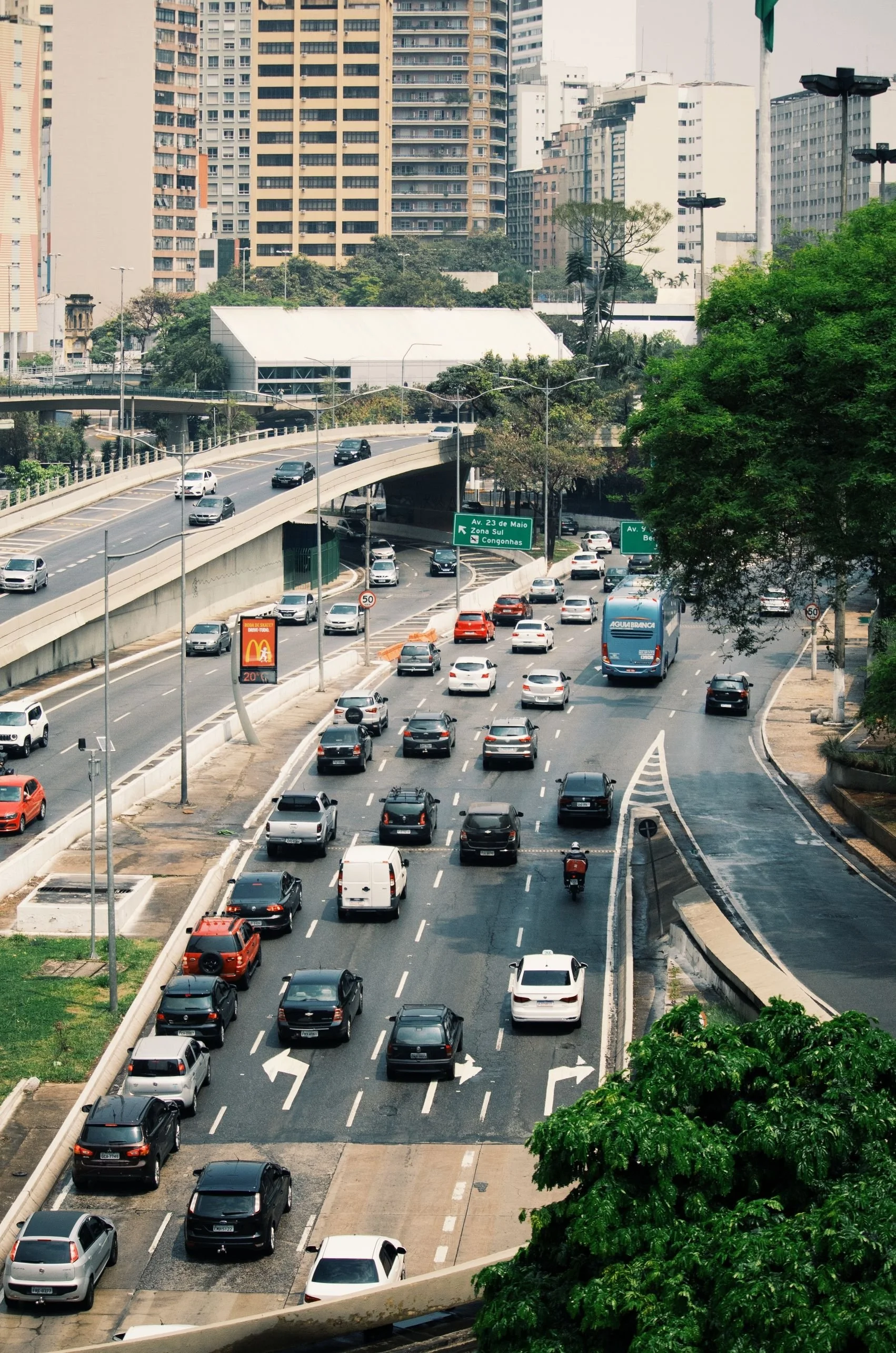 A city street with multiple lanes of traffic, several cars, and tall buildings in the background. There are green traffic signs, trees, and a McDonald's billboard.