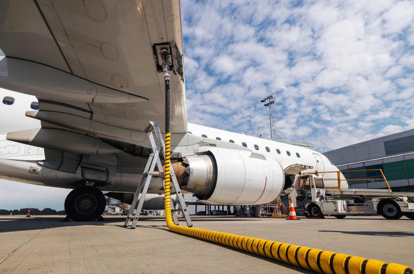 Close-up of aircraft ground maintenance, showing a jet engine, fuel hose, ladder, and service vehicle on tarmac at airport with terminal building and cloudy sky in background.