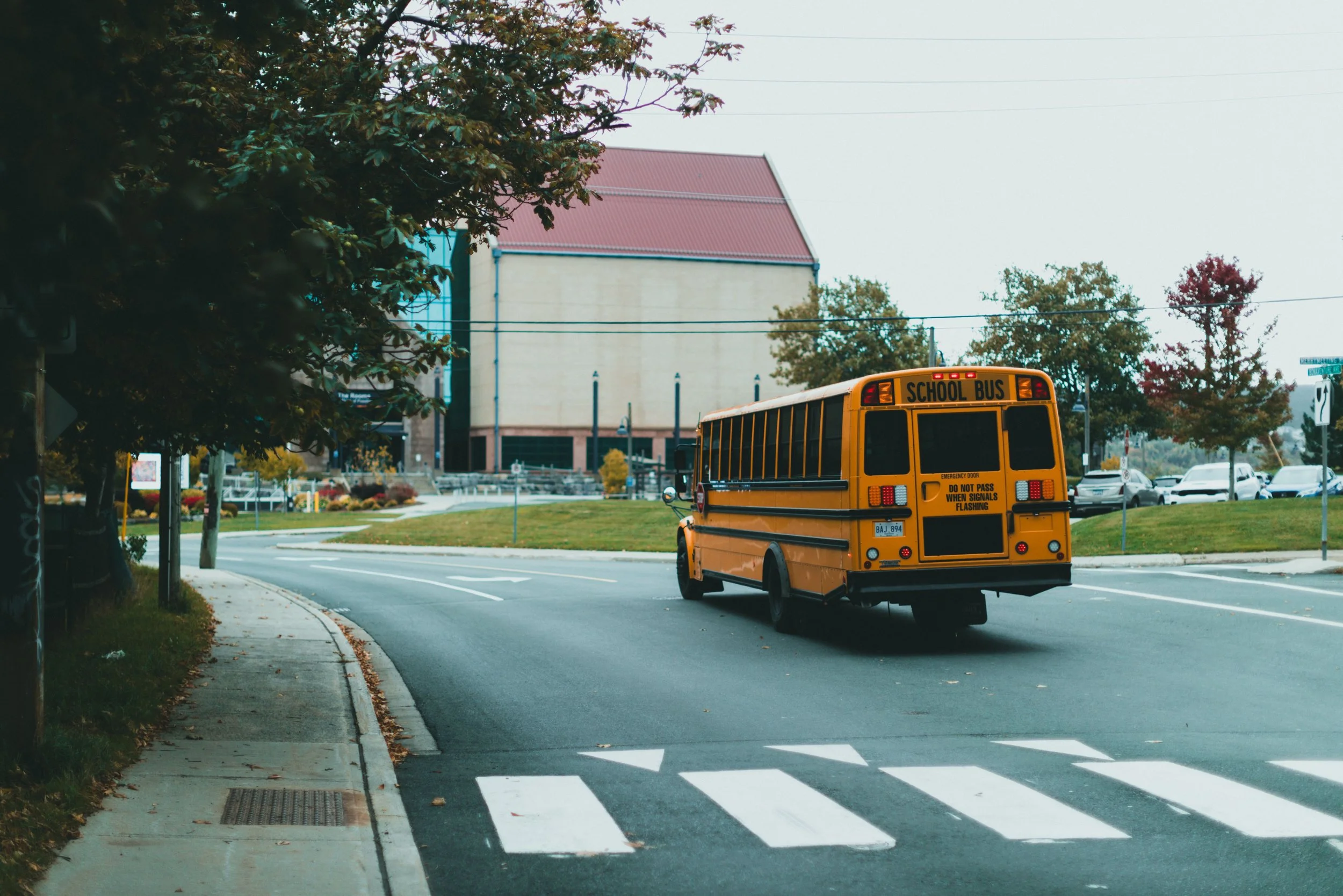A yellow school bus driving on a road near a school building with trees in the background.