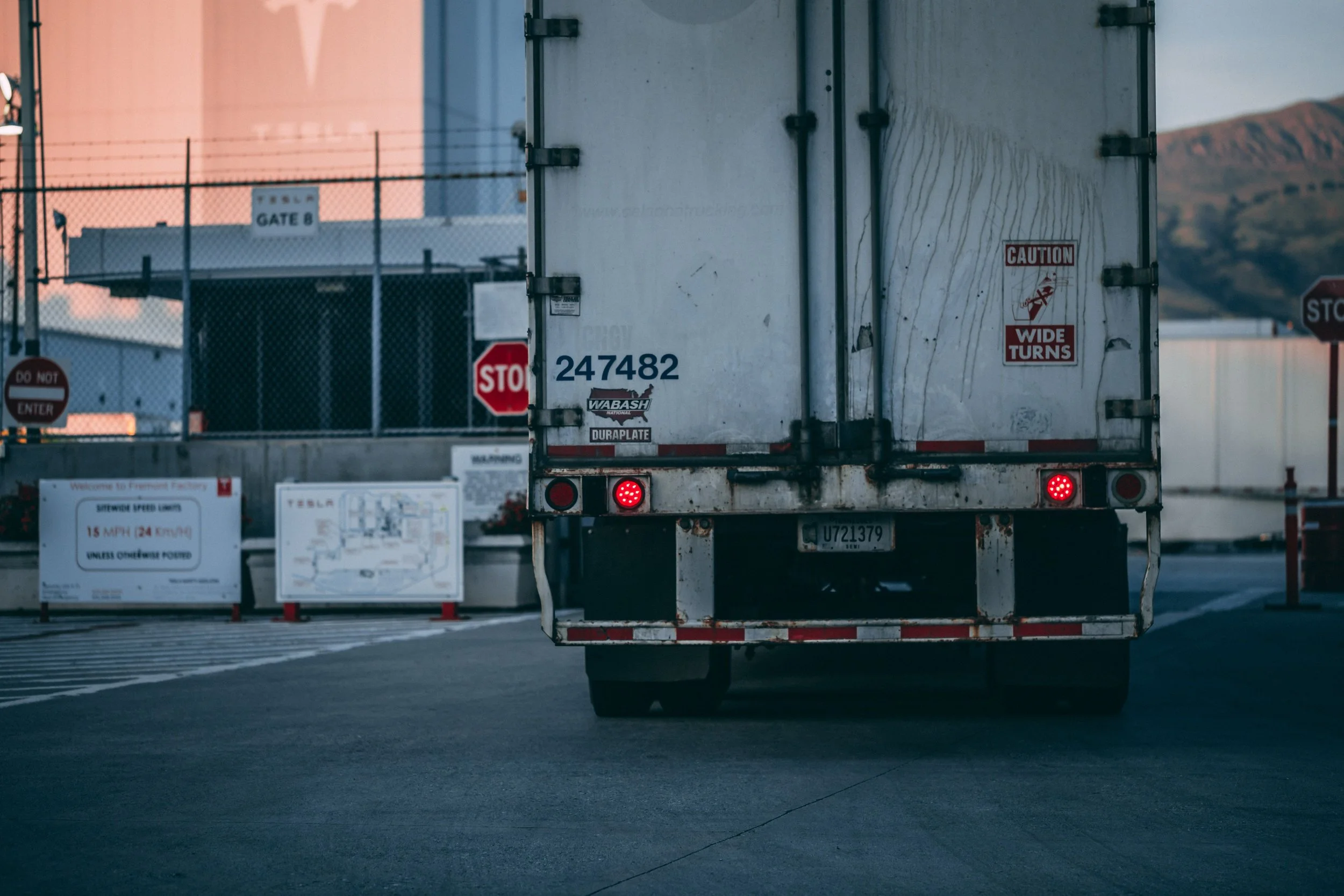 The back of a large white truck with warning signs, including 'Caution Wide Turns,' and various stickers and numbers. The truck is on a road, with a fencing area. The background features mountains and a clear sky at dusk.