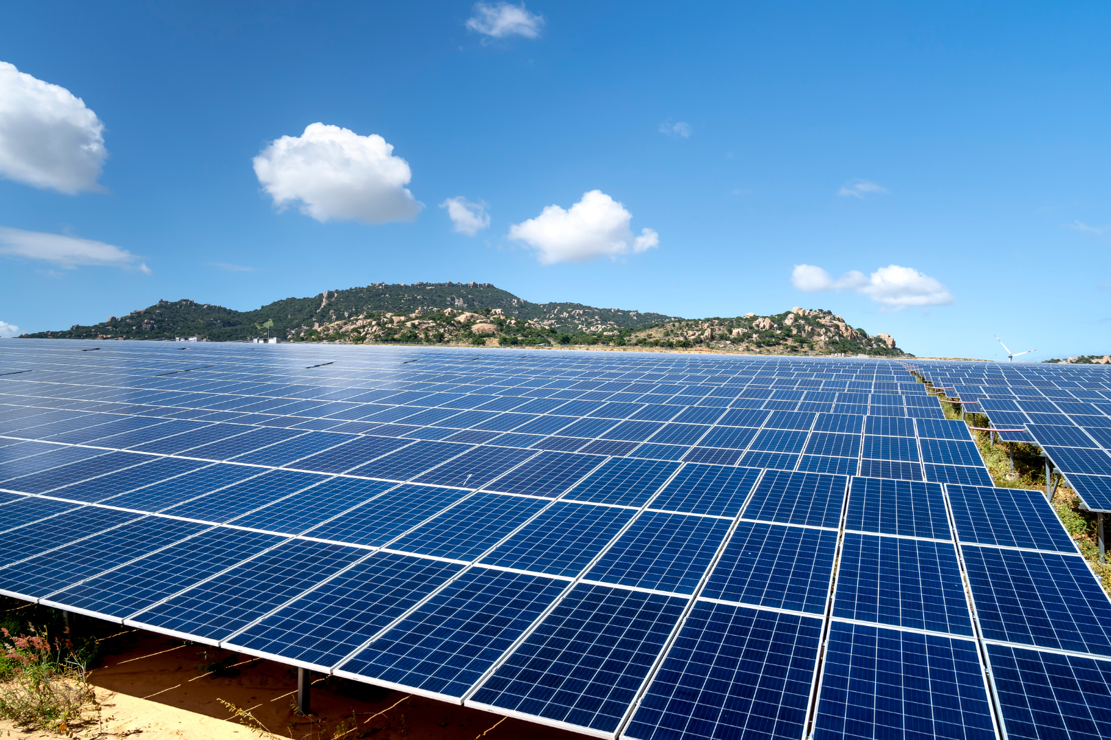 A large solar panel farm with numerous blue solar panels arranged in rows under a blue sky with scattered white clouds. In the background, there are green hills and a mountain.