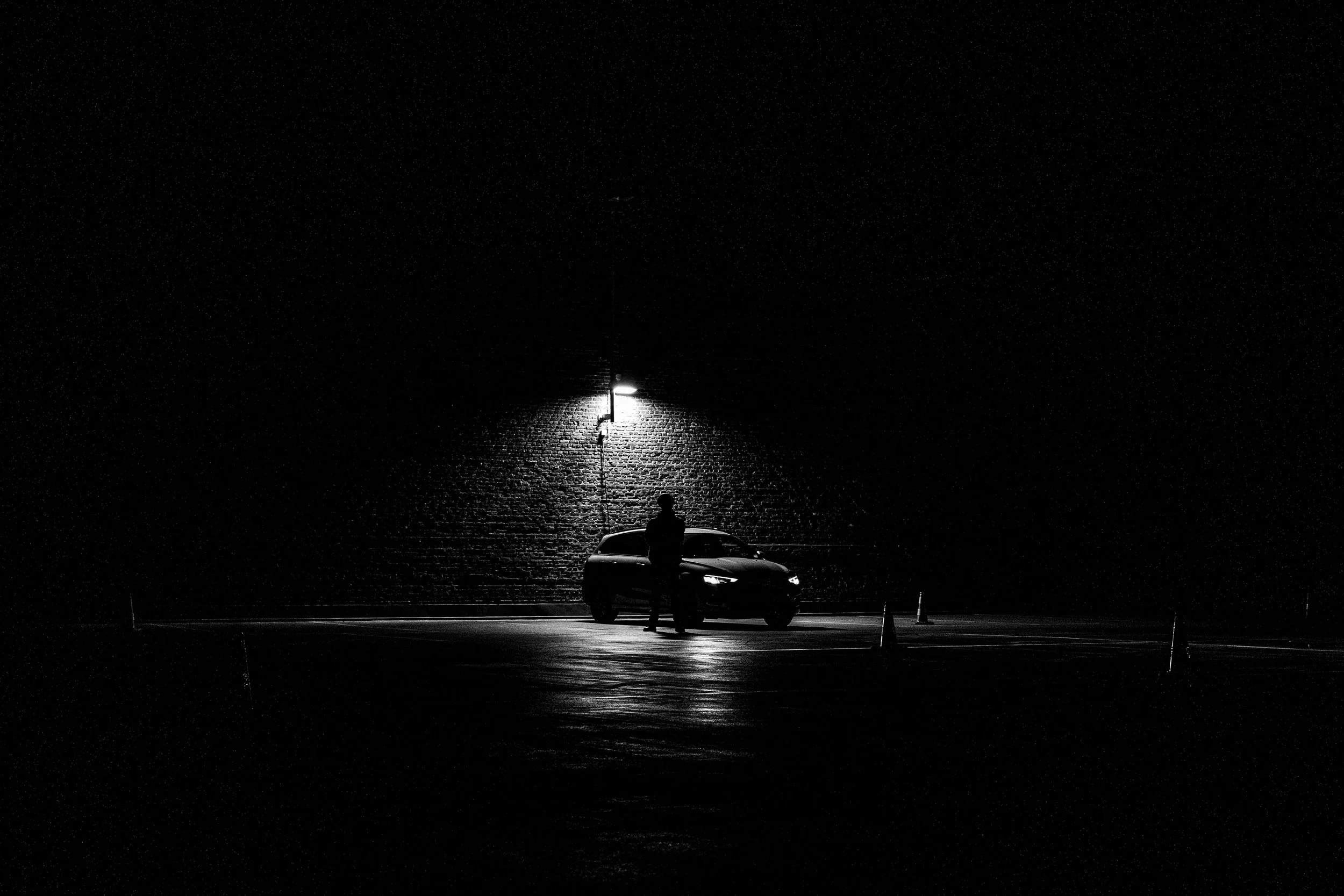 A black and white photo of a person standing next to a car at night, illuminated by a single streetlight against a dark brick wall background.