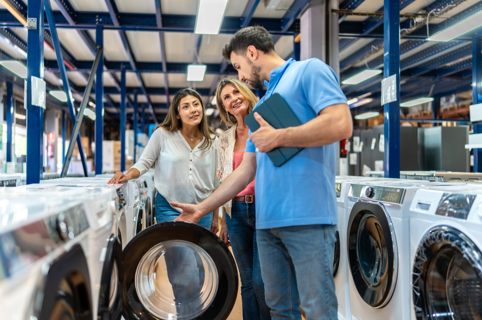 Two women shopping for washing machines in an appliance store, with a store employee explaining features to two women.