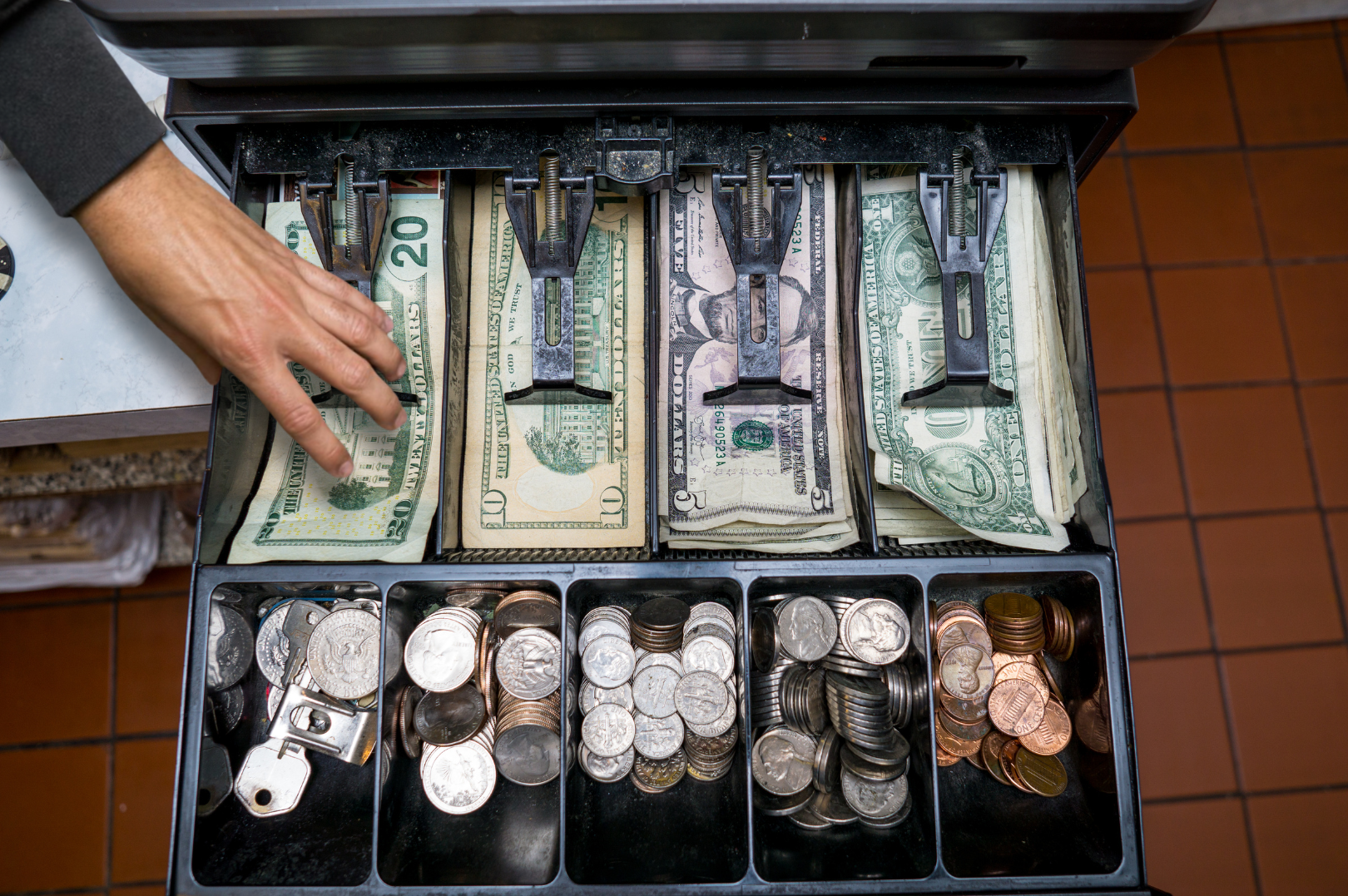 A cash register drawer open with various US dollar bills and coins organized in compartments, with a hand reaching over the counter into the cash register.