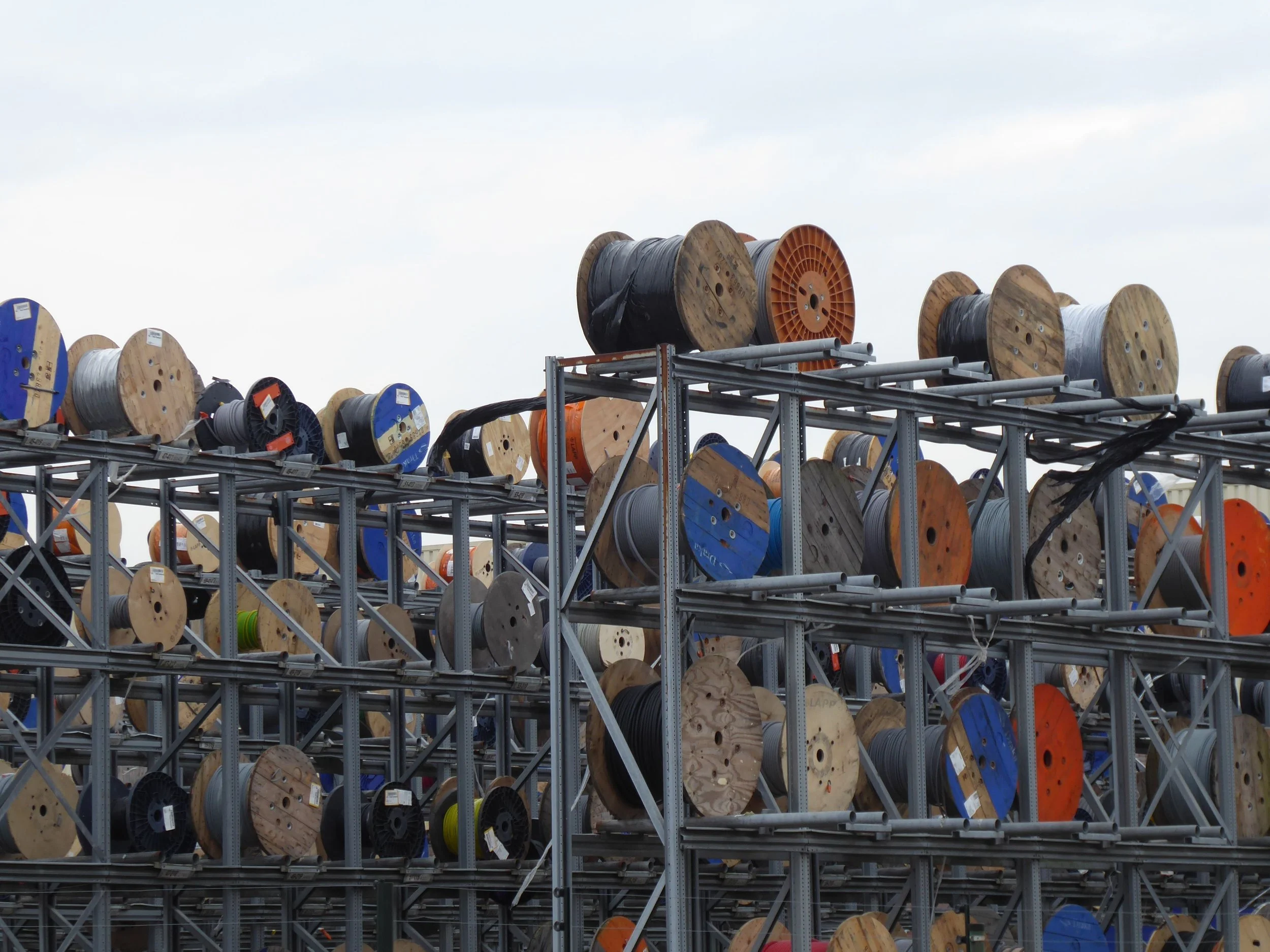 Several large spools of electrical wire stored on metal racks outdoors, with a cloudy sky in the background.