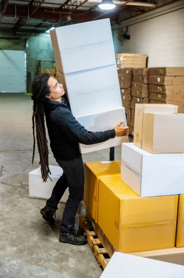 A woman with long dreadlocks wearing a black hoodie and pants, lifting a large box in a warehouse with stacks of boxes in the background.