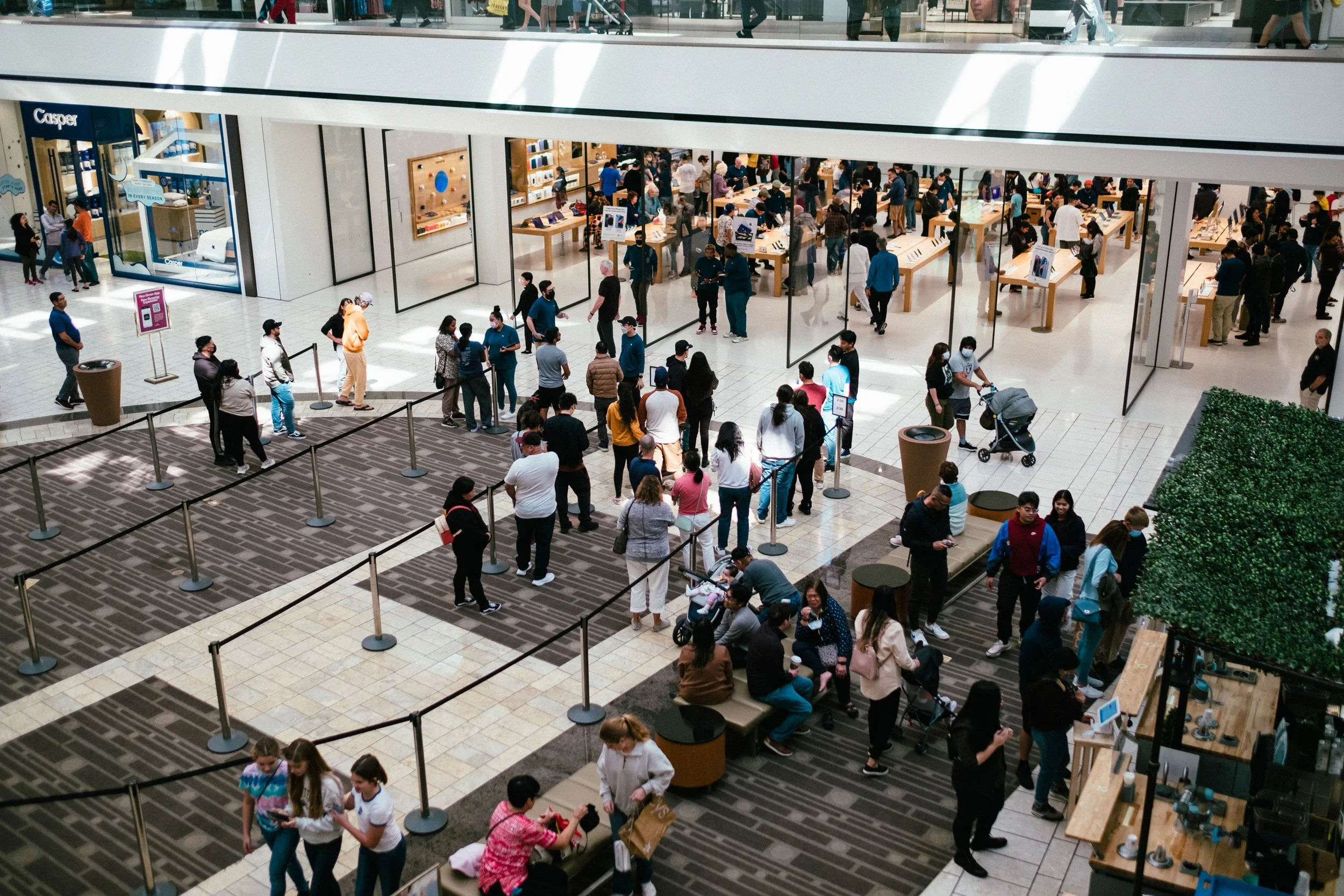 A busy indoor shopping mall with people walking, shopping, and standing in line. The lower level has several people waiting in line near a store called Casper, with others walking past or pushing strollers. The upper level has people browsing or walking.