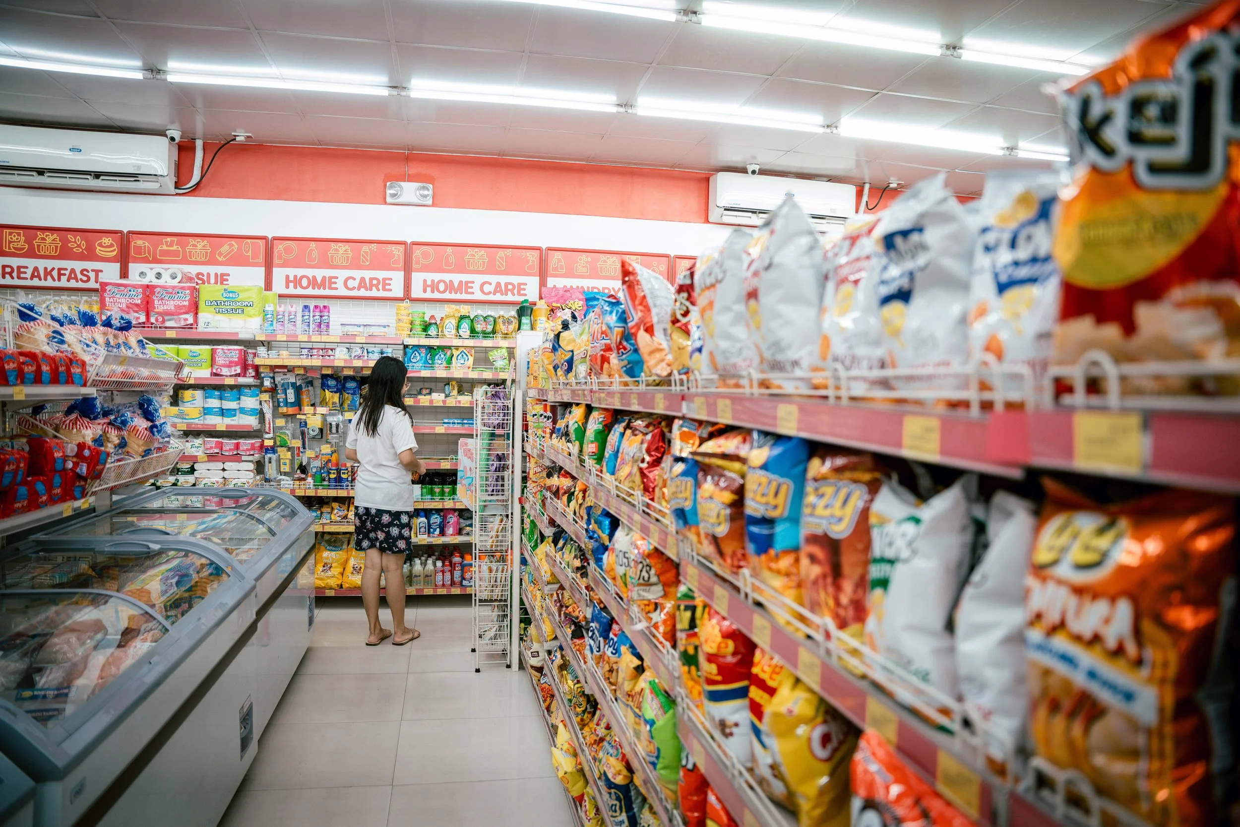 A woman shopping in a convenience store aisle filled with snack bags on the right and frozen foods on the left. Overhead signs indicate sections like breakfast and home care.