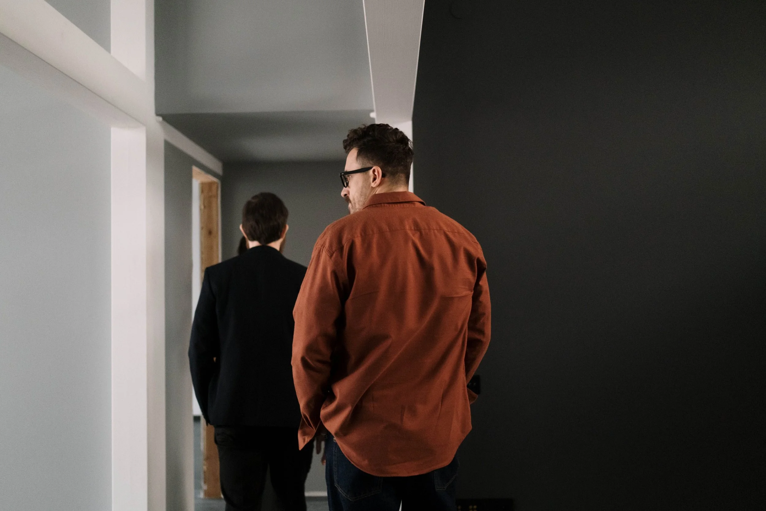 Two men walking down a hallway, one wearing a brown jacket and glasses, the other wearing a black jacket, with the back of their heads visible.