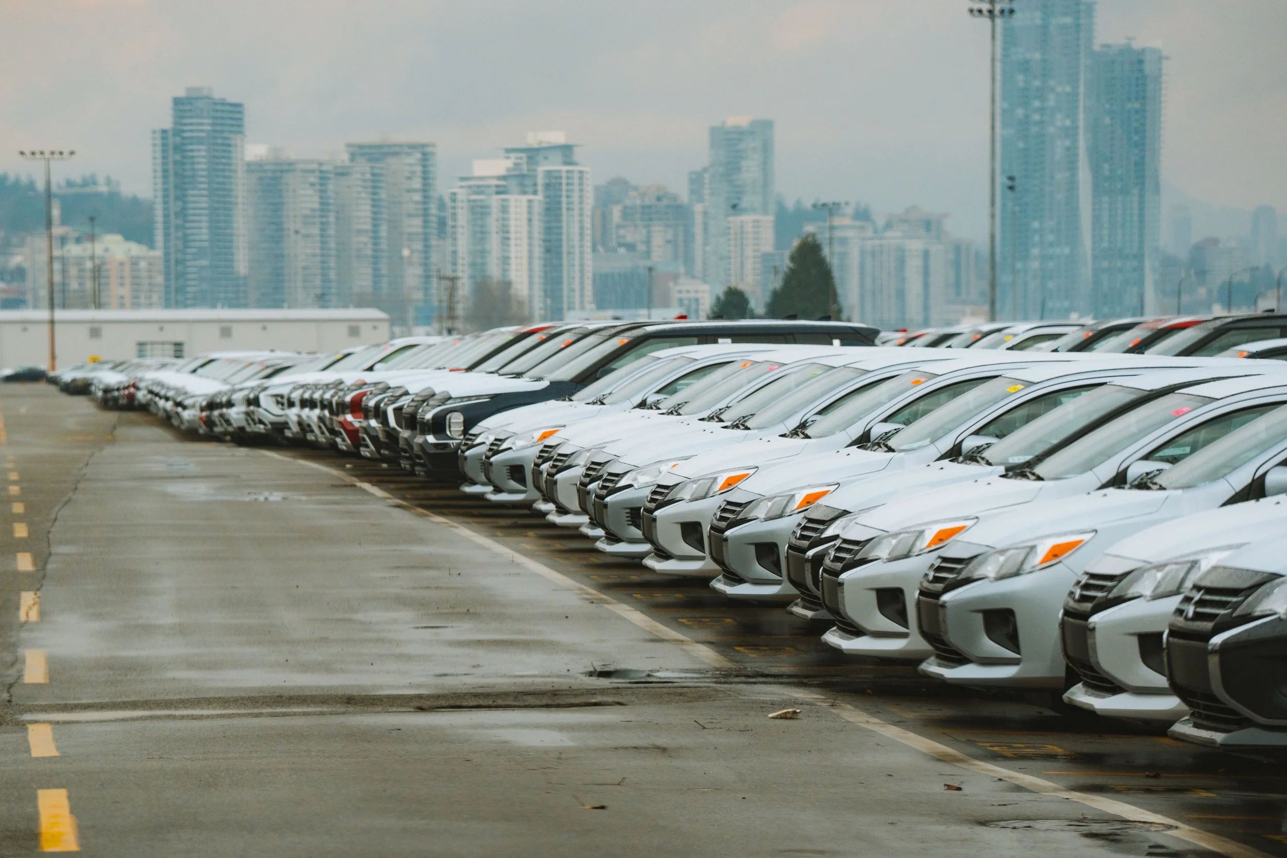 A large parking lot filled with rows of white cars, with a city skyline of tall buildings in the background overcast sky.