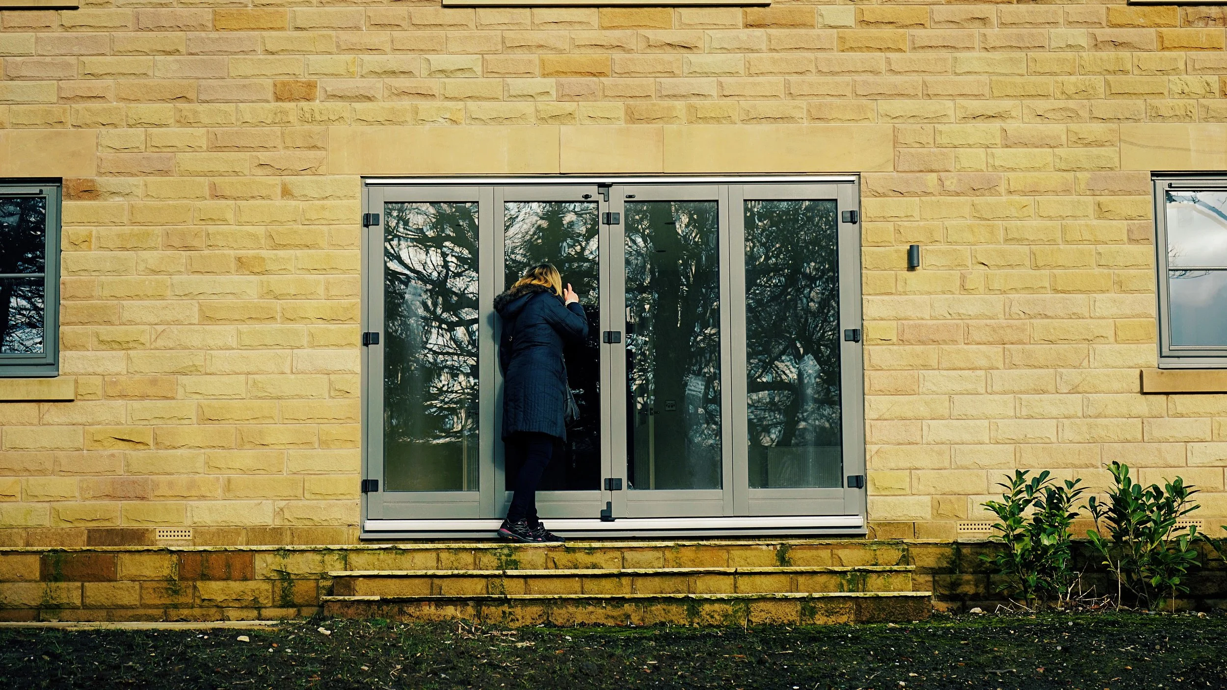 A woman with blonde hair, wearing a dark coat with a hood, is standing outside and looking through the glass door of a yellow brick building.