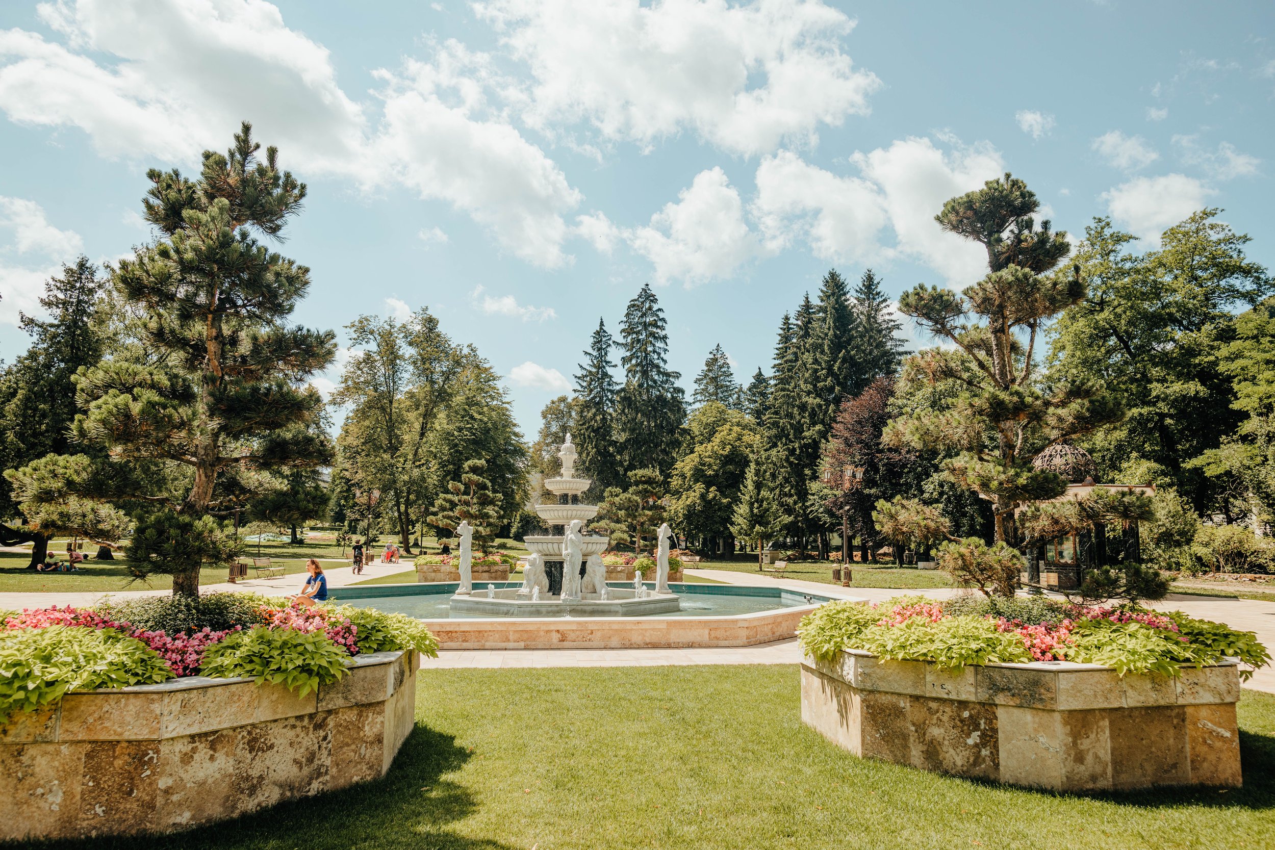 A public park with a decorative stone fountain surrounded by greenery and flowerbeds, tall pine trees, and a blue sky with clouds.