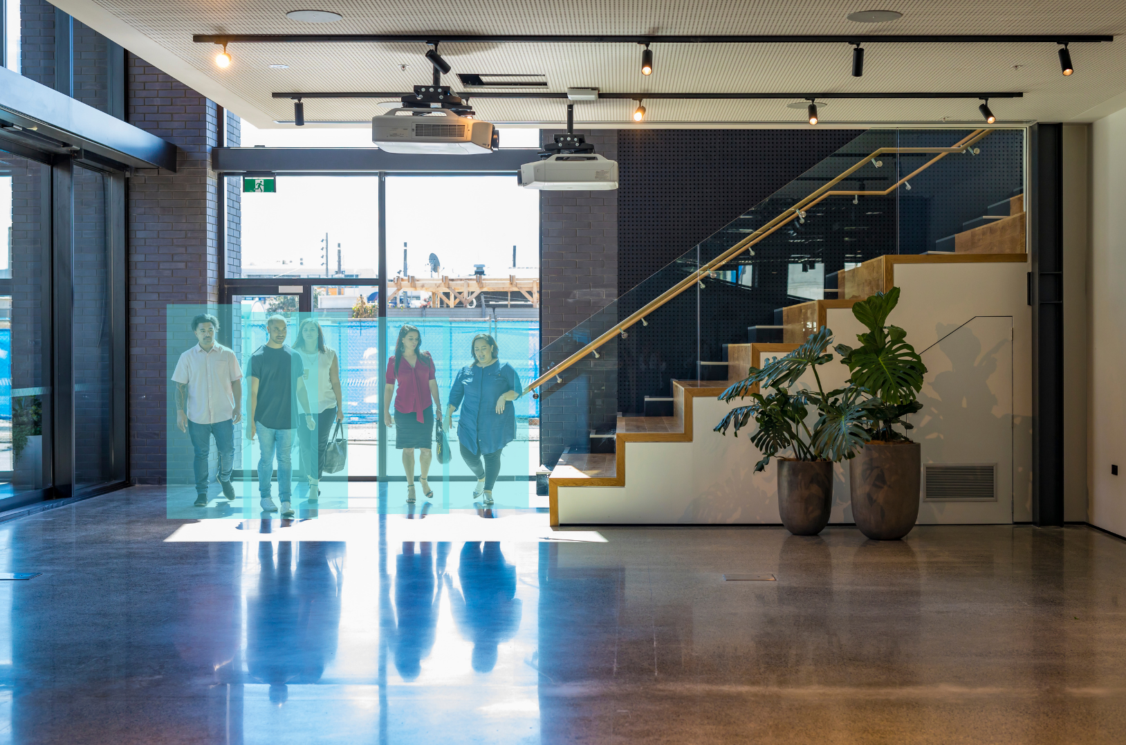 People walking through a modern indoor space with glass doors, a staircase with wooden steps and a glass railing, and large potted plants, with natural light coming through the windows.