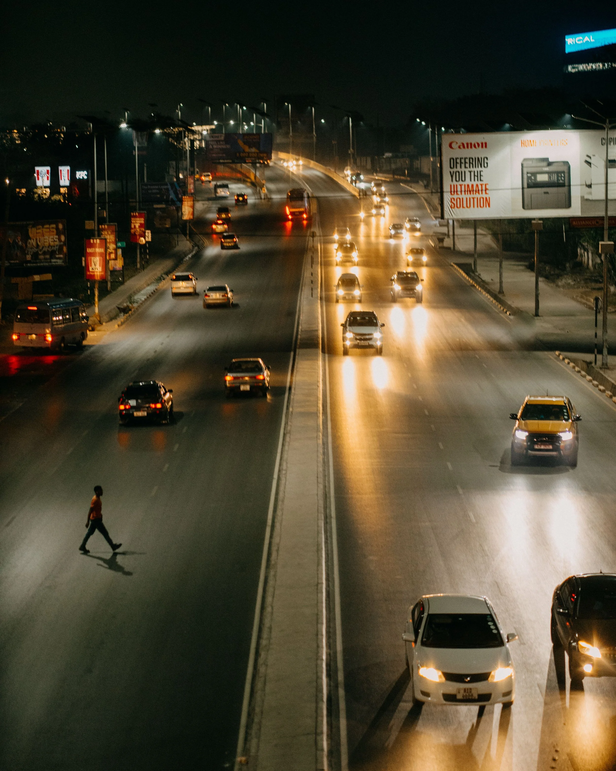City street at night with a pedestrian walking across, cars on the road with headlights on, illuminated billboards, and a large billboard on the right advertising a Canon product.