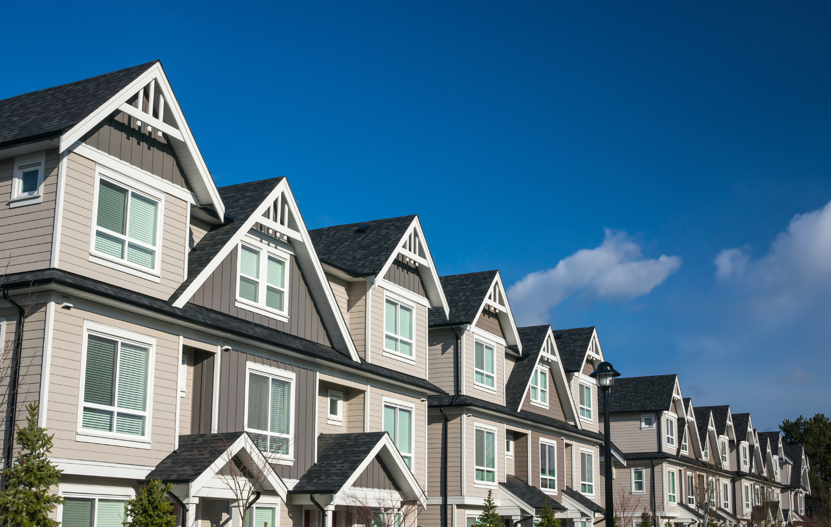 A row of modern townhouse apartments with beige and dark brown siding, white trim, and gabled roofs under a blue sky with a few clouds.