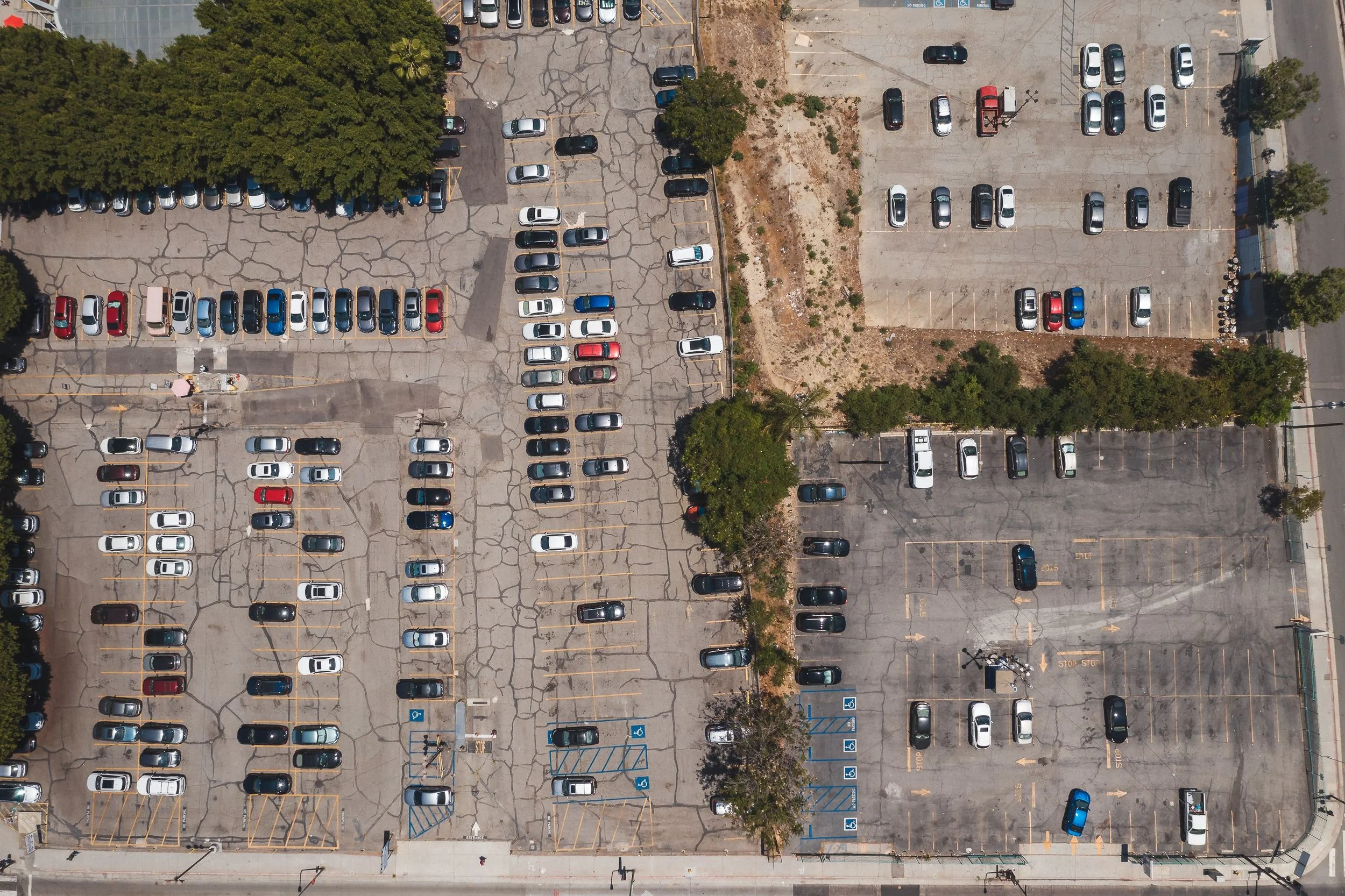 An aerial view of a parking lot with scattered cars, trees, and some empty parking spaces. The lot shows areas with different levels of wear and tear, and some designated parking spots for handicapped.