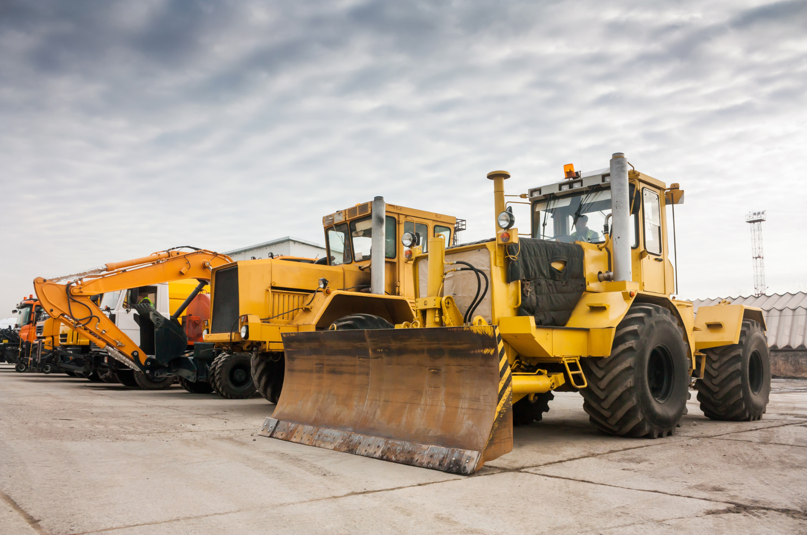 A row of yellow construction vehicles, including a bulldozer, parked on a concrete surface outdoors under a cloudy sky.