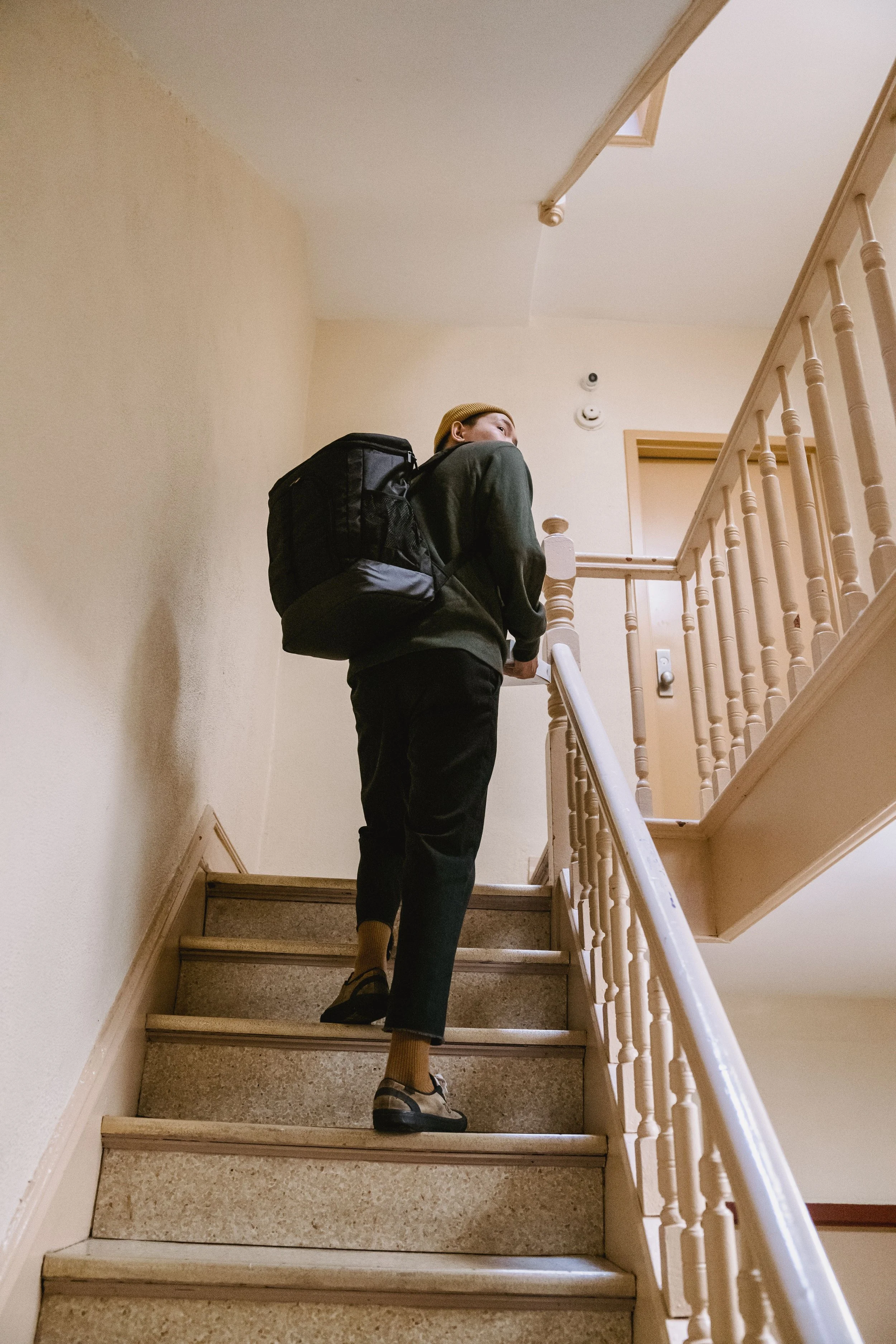 Person with a backpack and yellow beanie walking up a staircase inside a building, with beige walls and wooden handrails.
