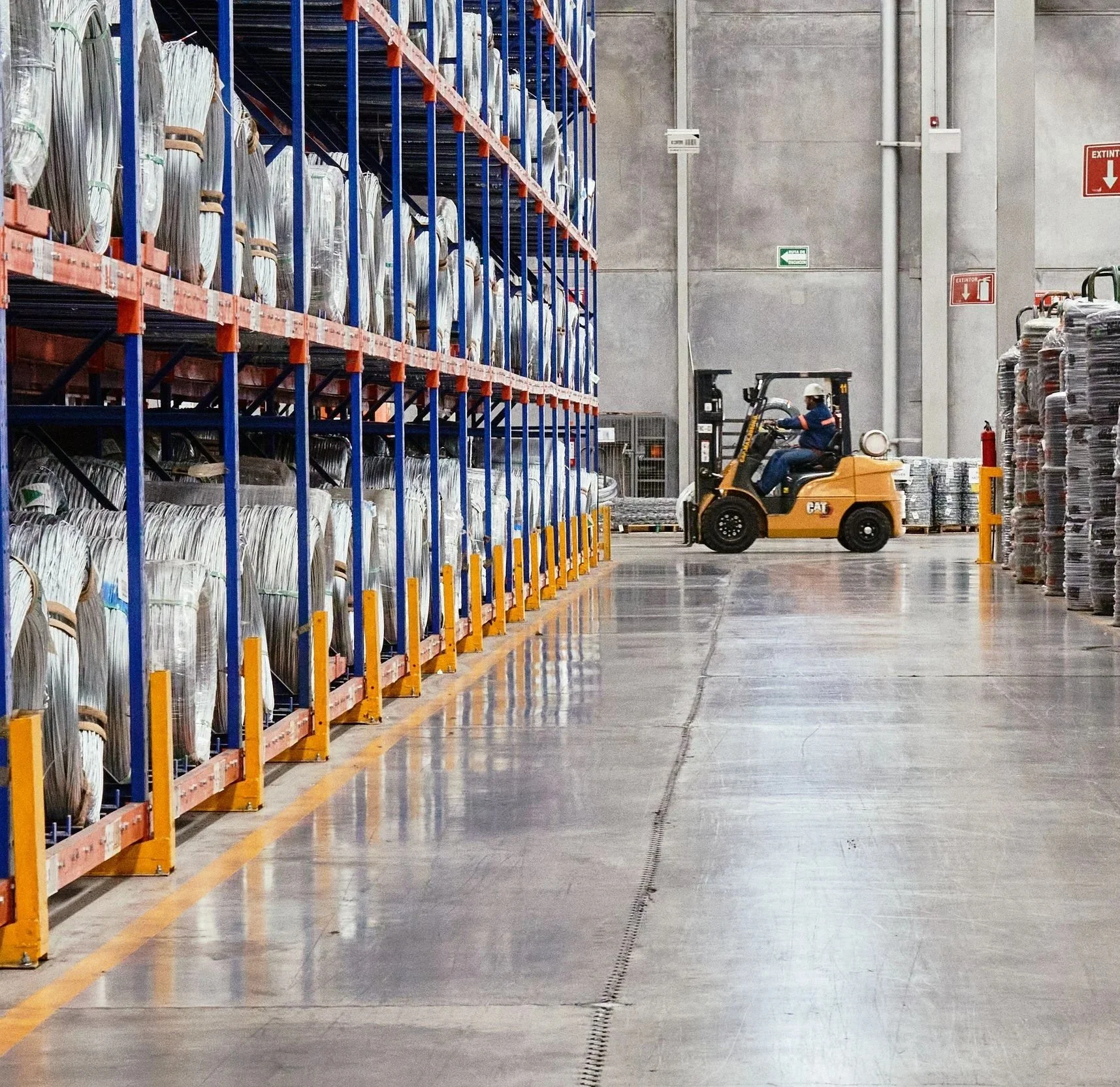 Warehouse interior with tall shelving units stacked with coils of cable or wire, and a forklift operated by a worker in safety gear moving through the aisle.