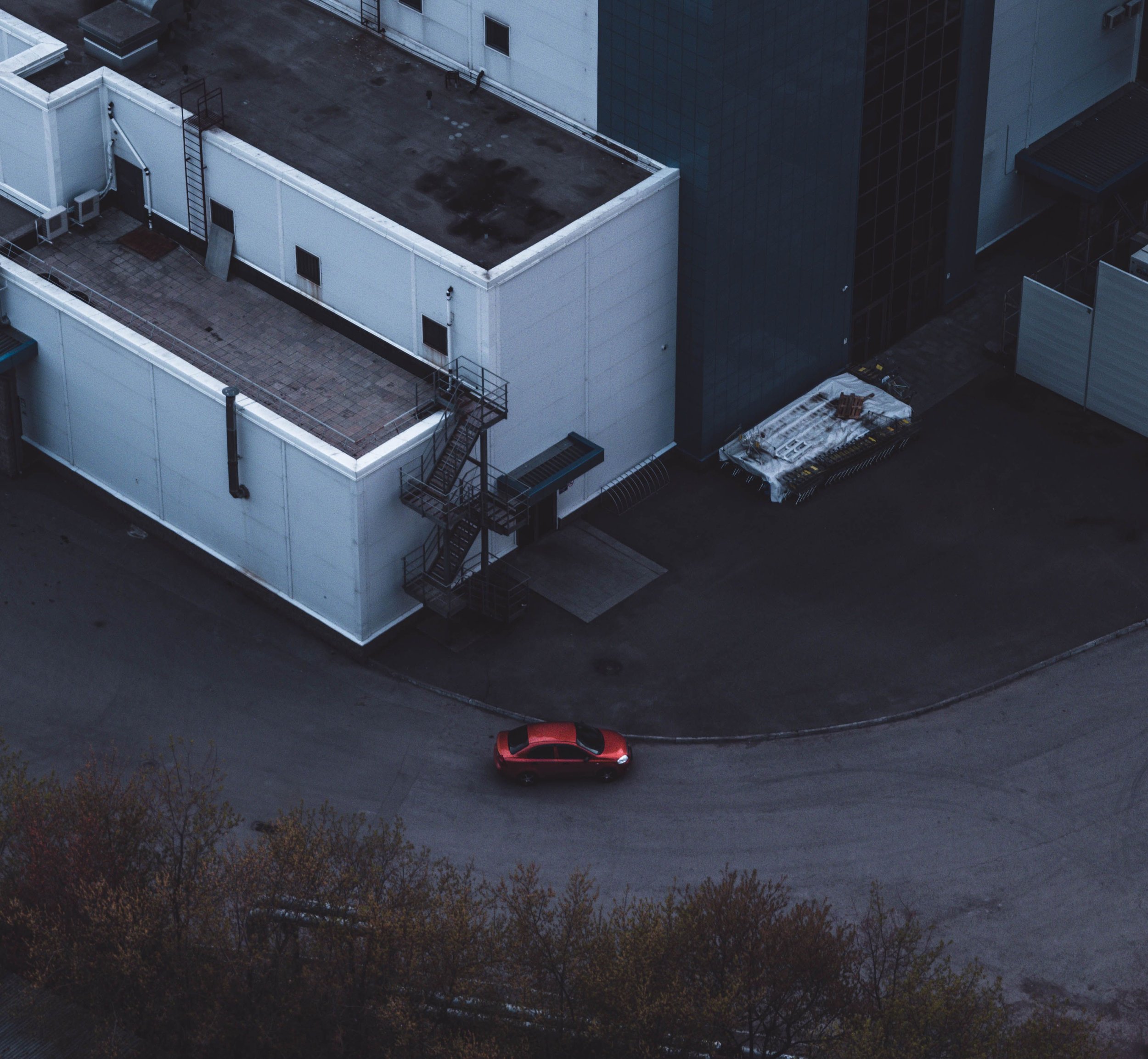 An aerial view of a white commercial building with a flat roof, a small rooftop area, and an external metal staircase. A red car is parked on the street below near some trees.