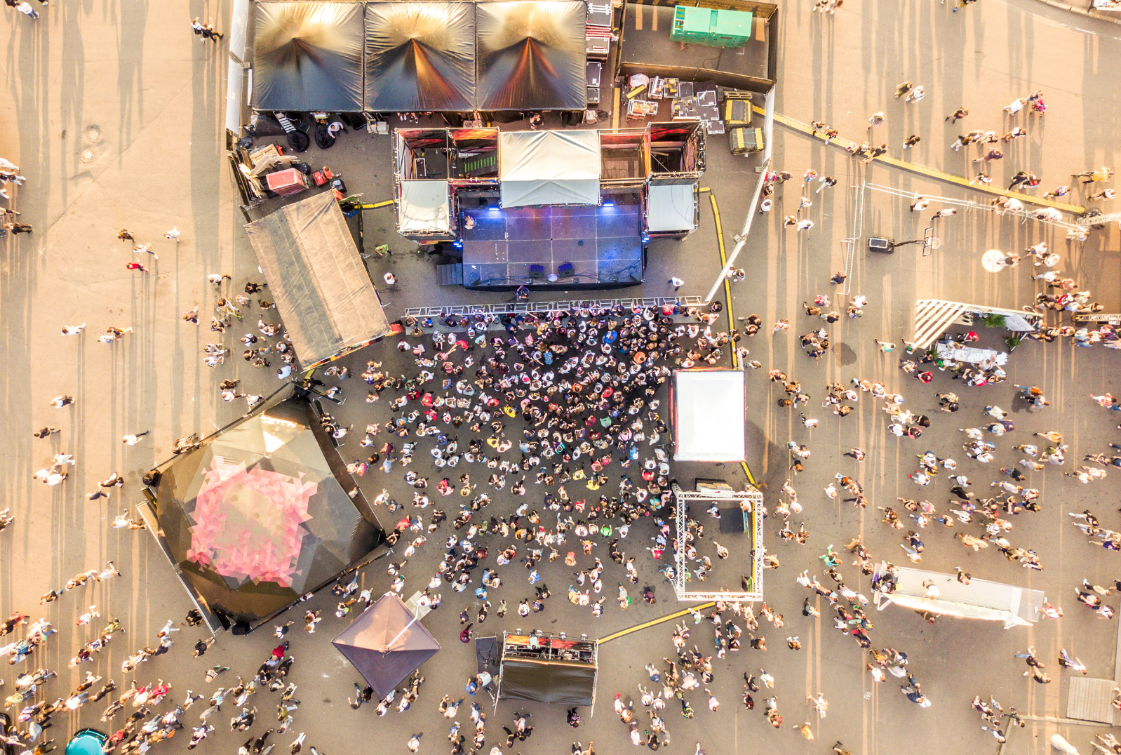 An aerial view of a crowded outdoor concert stage and audience on a city street during daylight, with tents, equipment, and people scattered around.
