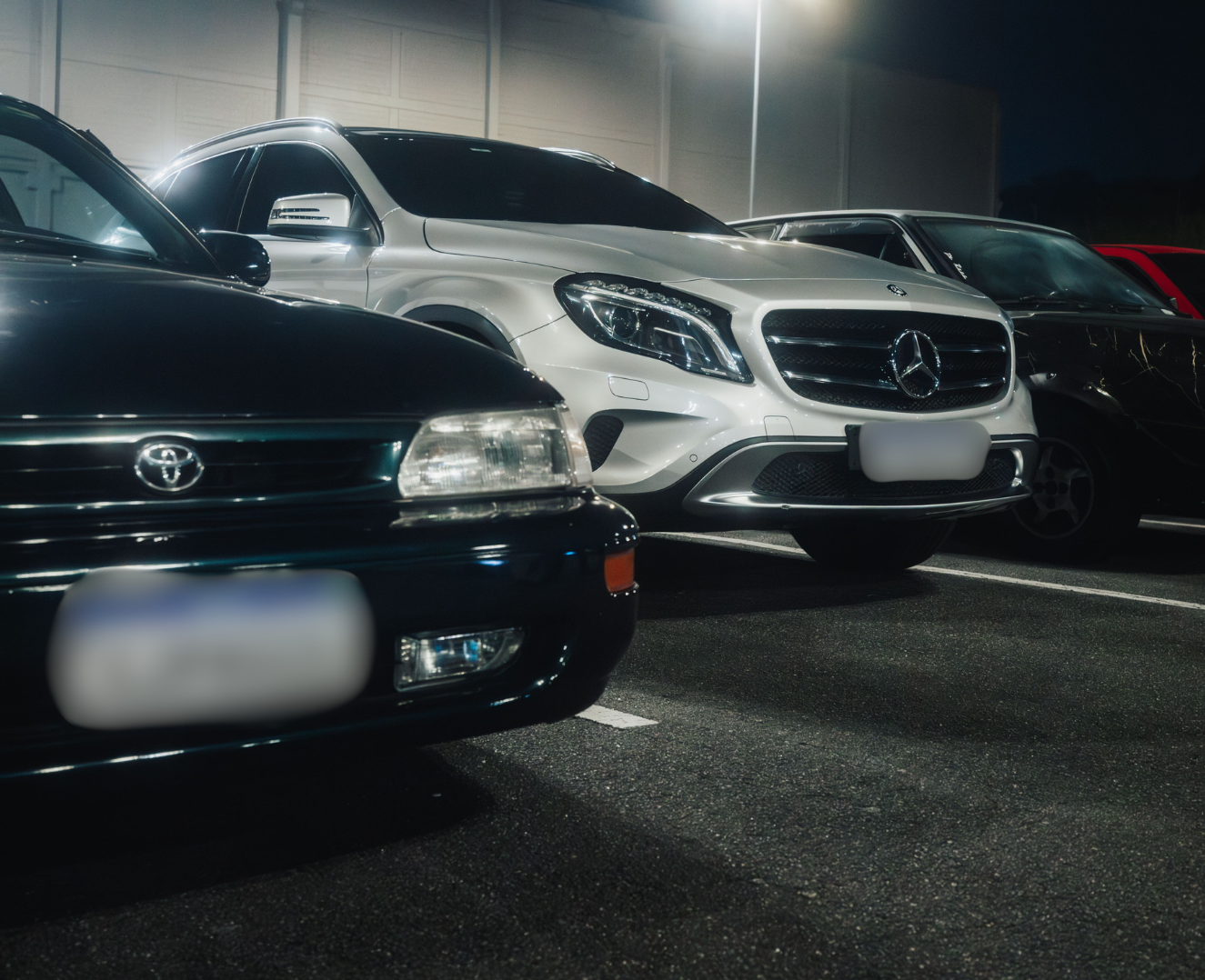 Various parked cars in a dimly lit parking lot at night, including a black Toyota, a silver Mercedes-Benz, and a partially visible red vehicle.