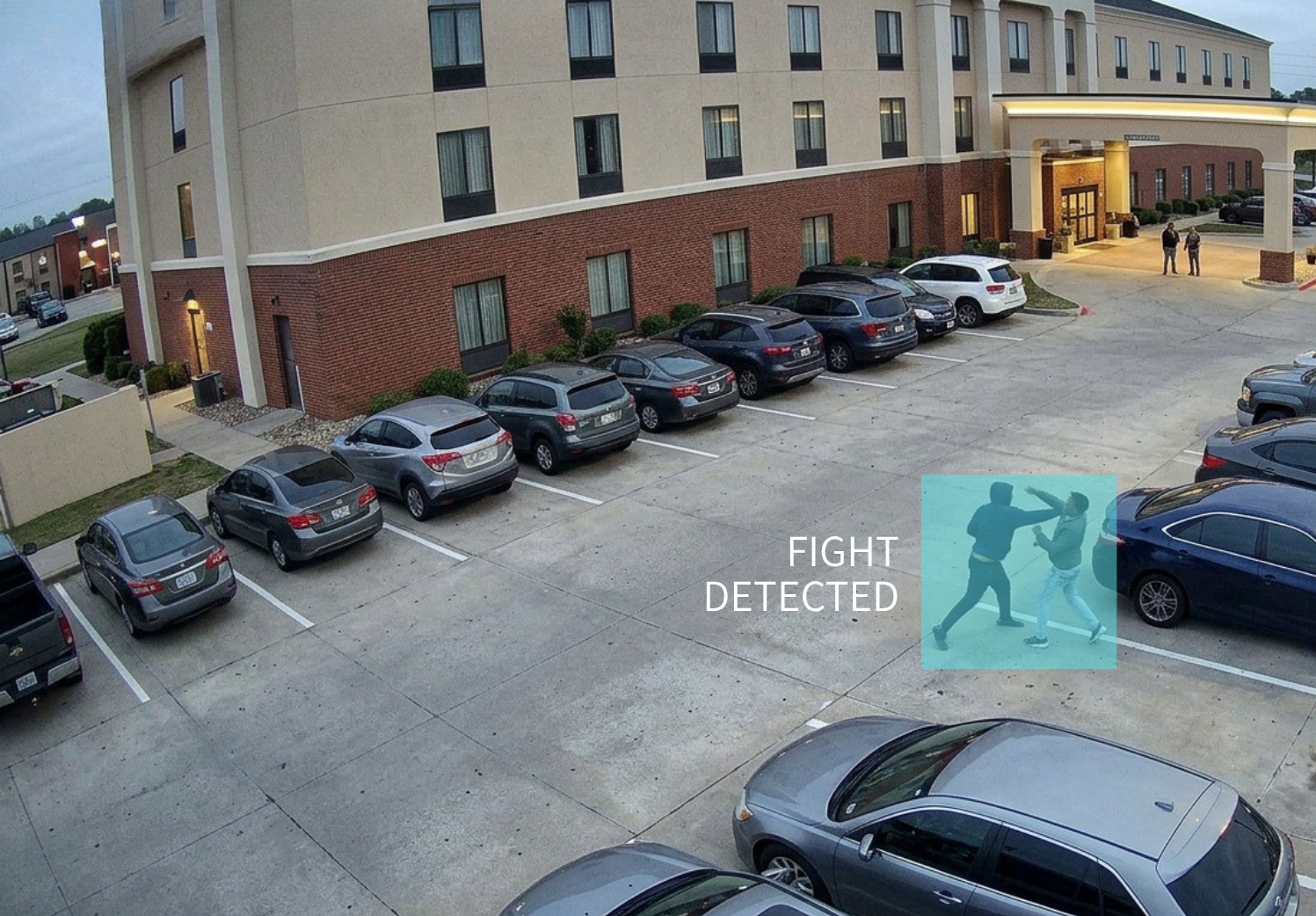 Two individuals engaged in a physical altercation in a parking lot outside a multi-story hotel building, with the words 'FIGHT DETECTED' overlaid on the image.