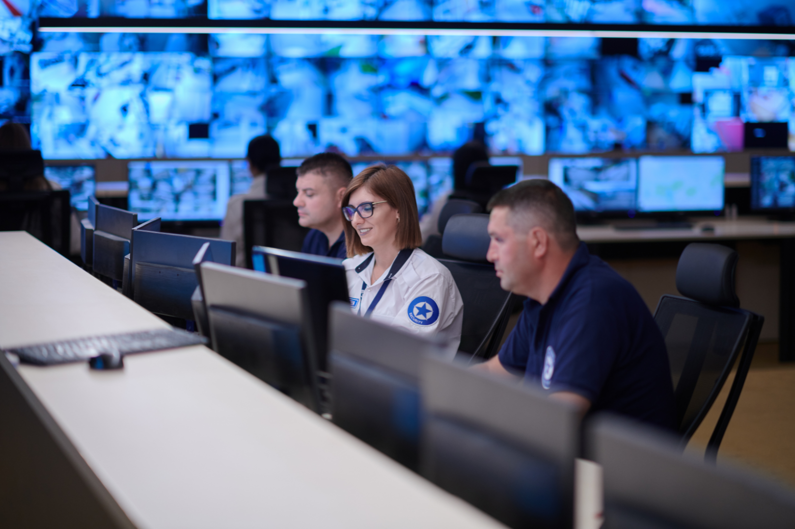 People working in a control room with multiple monitors showing surveillance footage, with three people focused on their screens in the foreground.