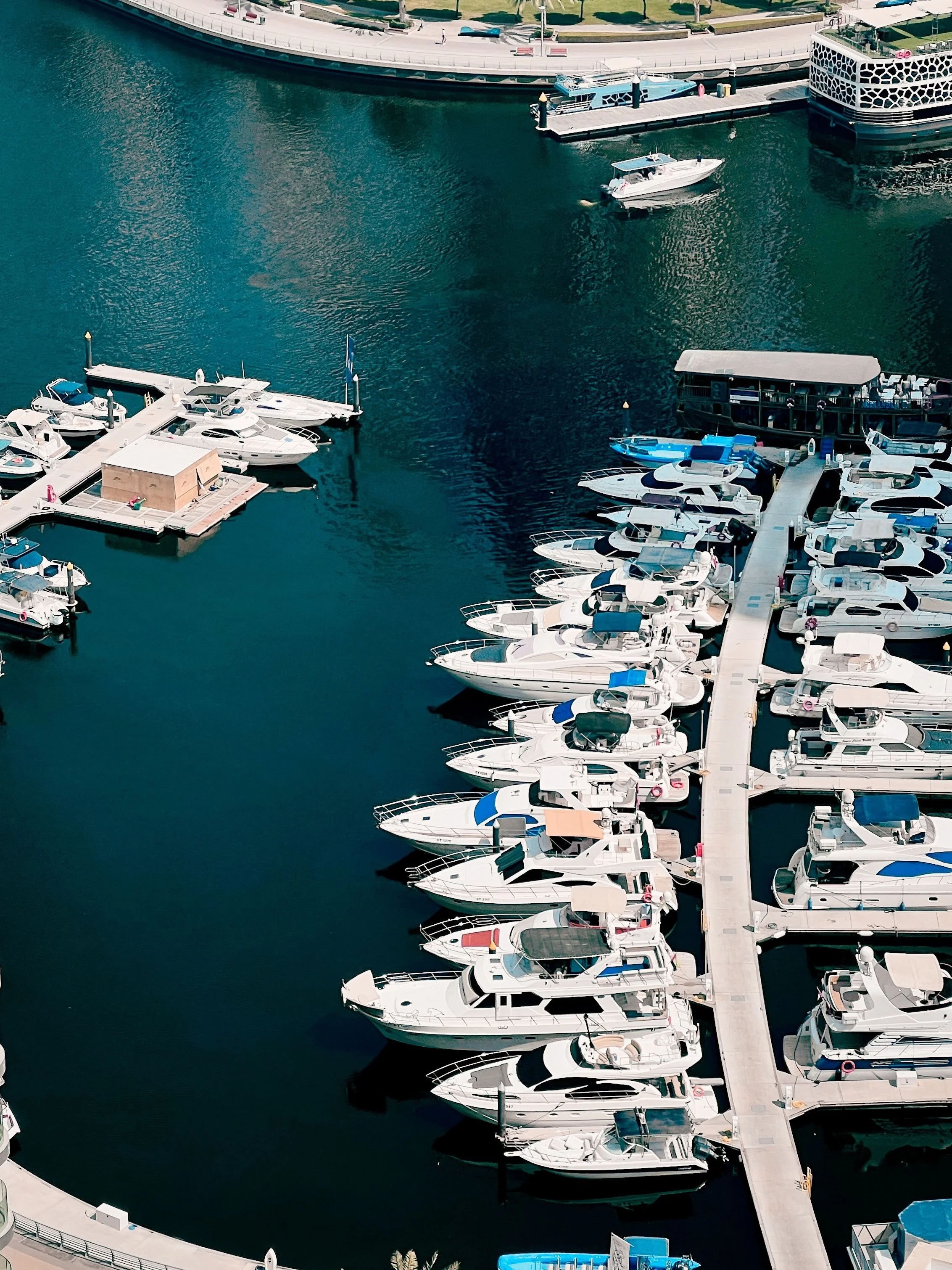 Aerial view of a marina with multiple docked yachts and boats, adjacent to a curved waterfront walkway and a modern building with a geometric facade.