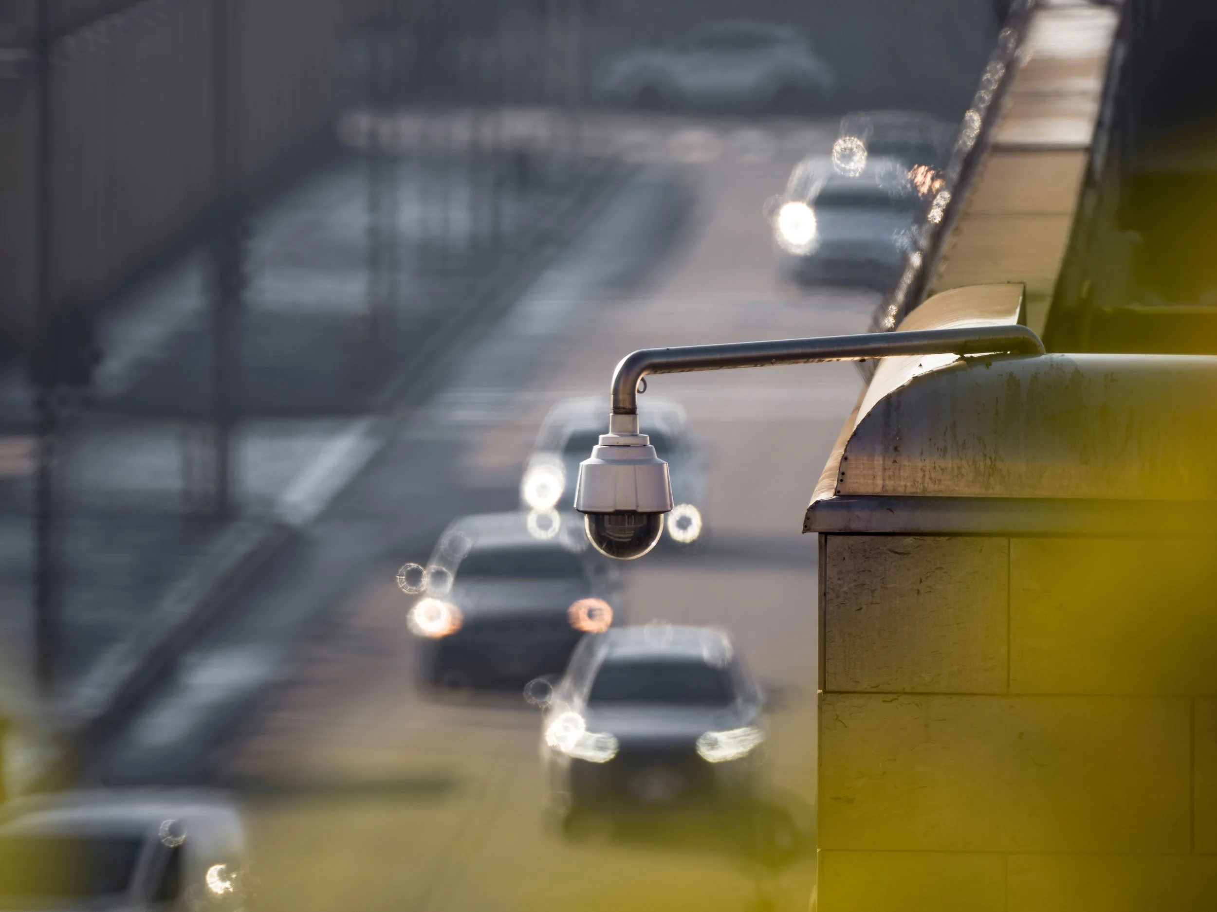 A surveillance camera mounted on a building structure overlooking a road entrance with multiple cars driving into the property.