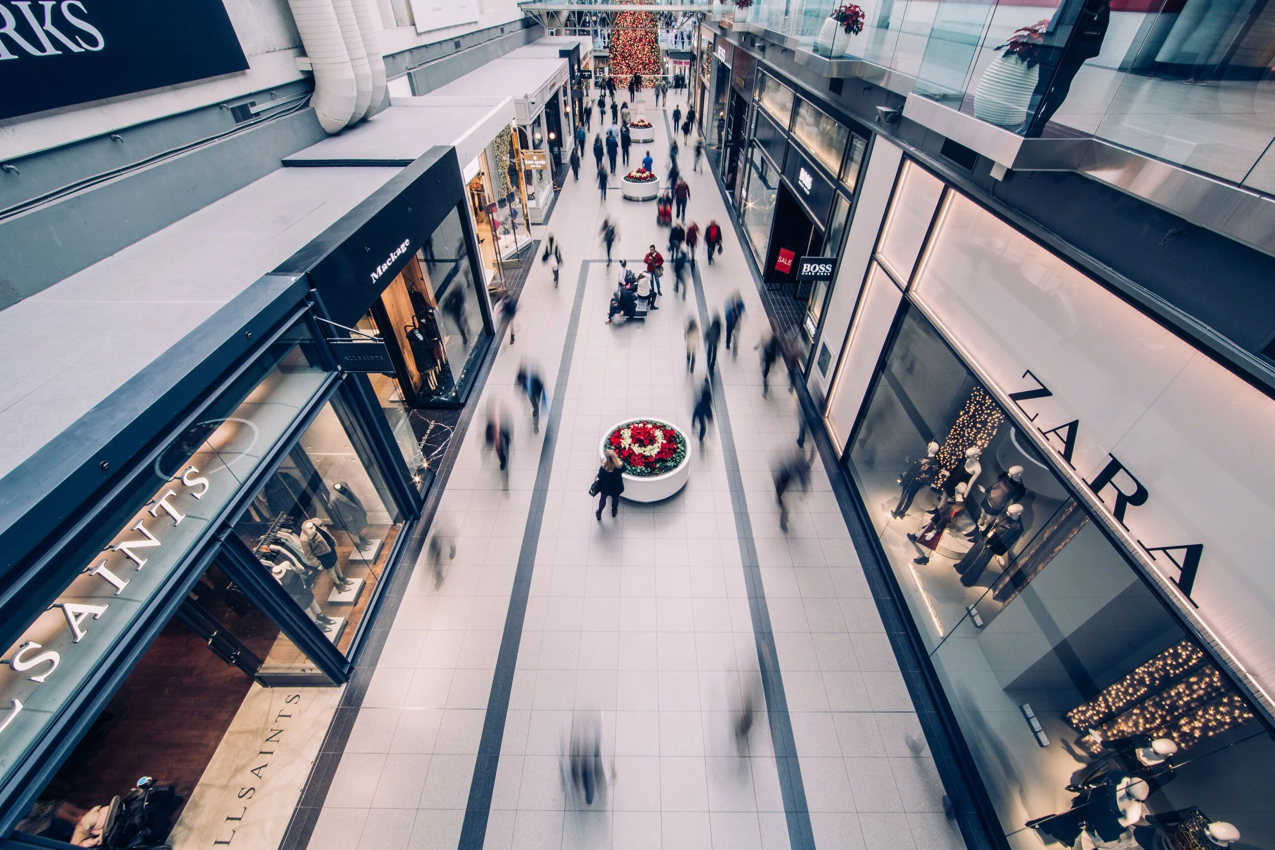 Interior view of a shopping mall with people walking and shopping, holiday decorations including a large Christmas tree and festive flower arrangements.