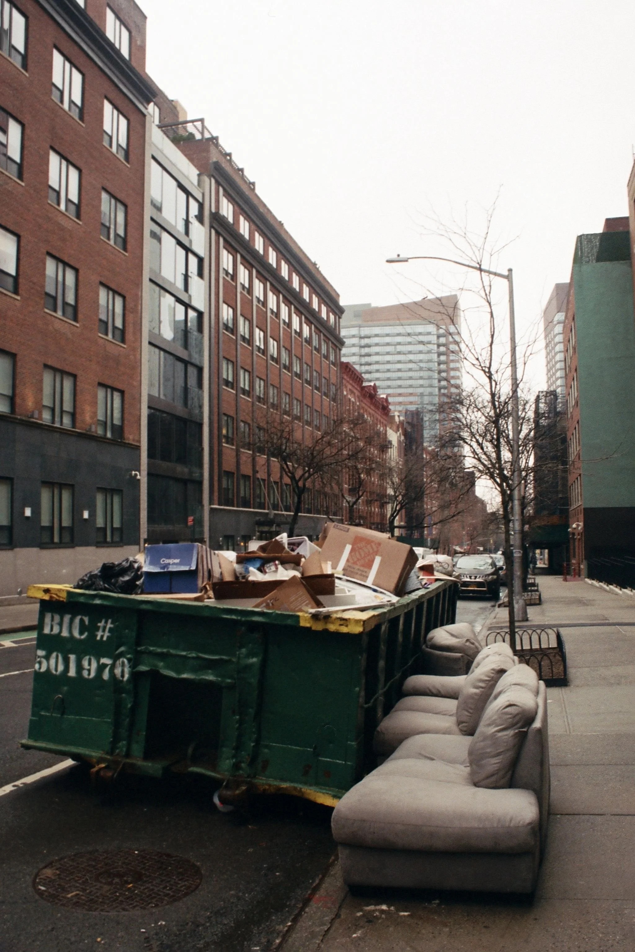 Street scene with a green dumpster filled with cardboard boxes and trash, a row of large beige sofas along the sidewalk, leafless trees, and multi-story brick and concrete buildings in the background.