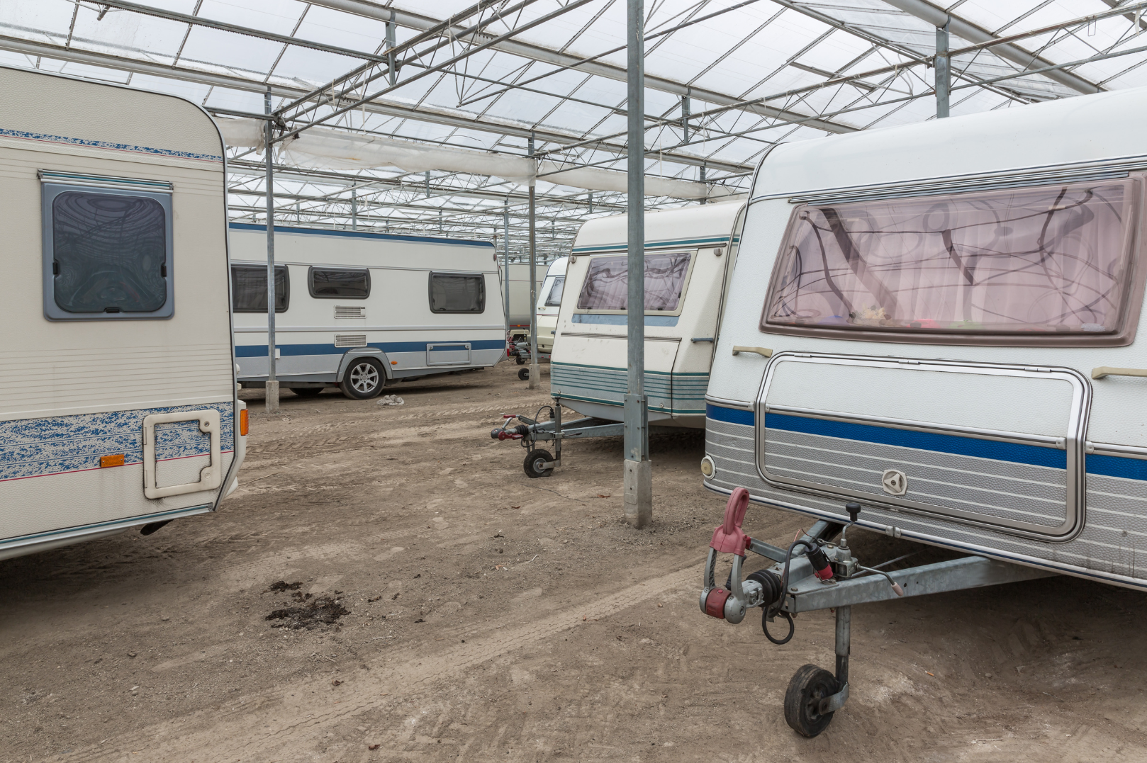Several travel trailers parked inside a large, partially transparent roofed structure. The trailers are of various sizes with some showing windows and doors. The ground is dirt, and some trailers have visible hitch attachments.