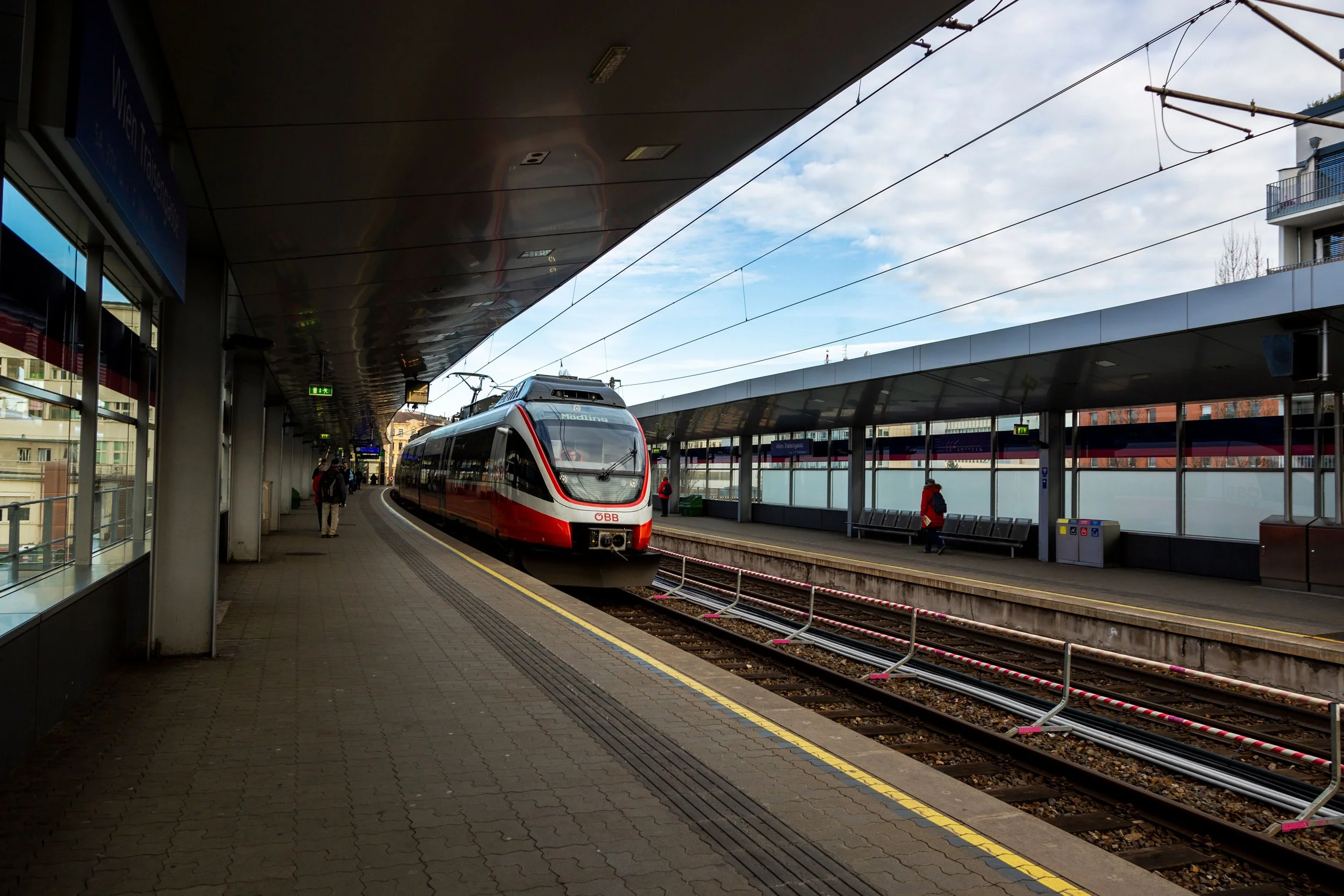 Interior of a train station with a modern white and red train on the platform, passengers waiting and walking, overhead electrical wiring, and large windows with a view of buildings outside.