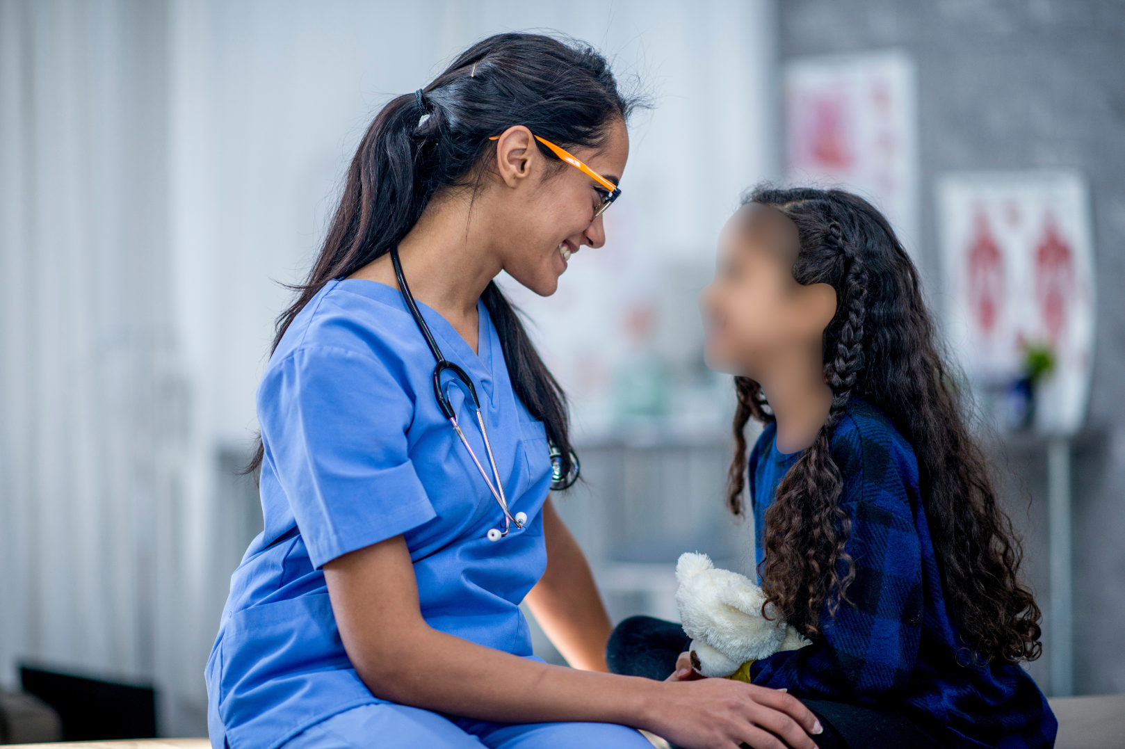 A female healthcare worker in blue scrubs and glasses smiling at a young girl with long curly hair, in a medical setting with posters in the background. The girls face is blurred using privacy ai.