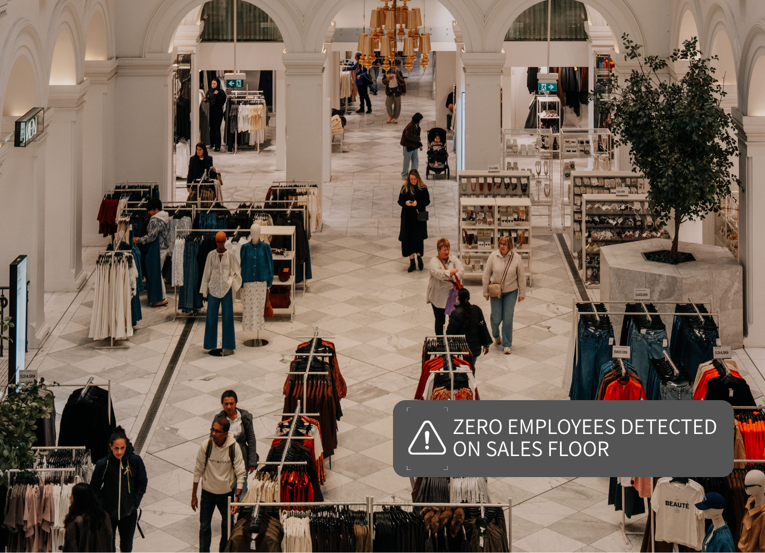Interior of a shopping mall with clothing displays and shoppers walking around, with a sign indicating zero employees detected on the sales floor.