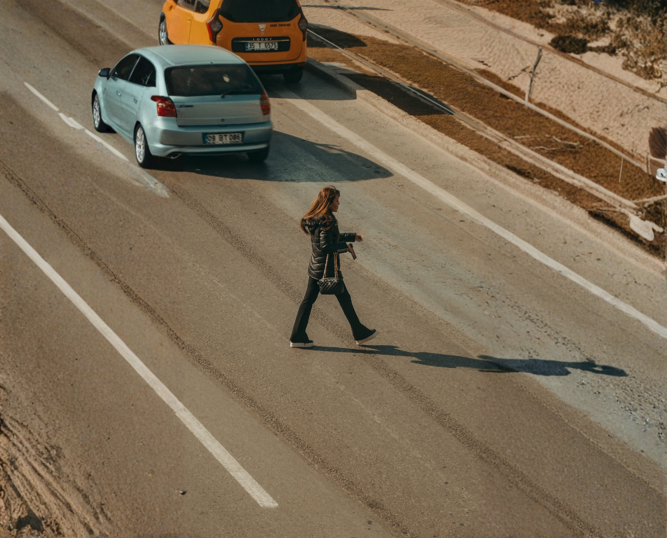 A woman walking across a city street with cars in the background, casting a long shadow on the road.