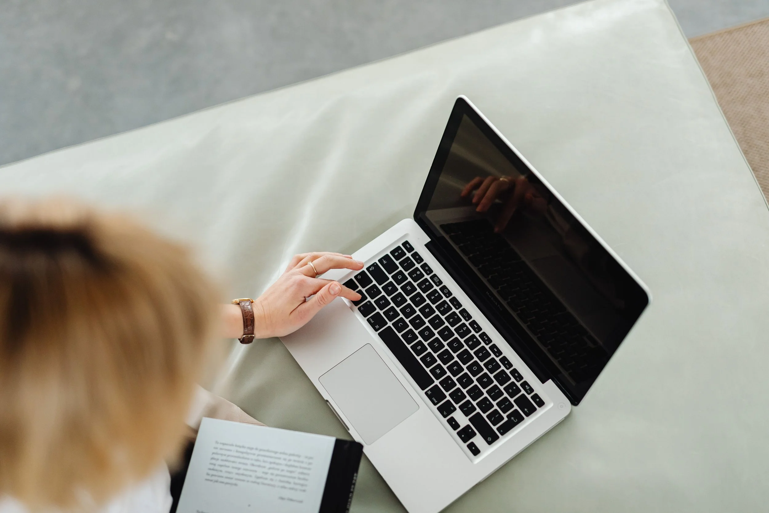 A person is working on a silver laptop at a white table, with an open book and a paper next to it. The person has a bracelet on their wrist and is in a room with neutral-colored furniture.