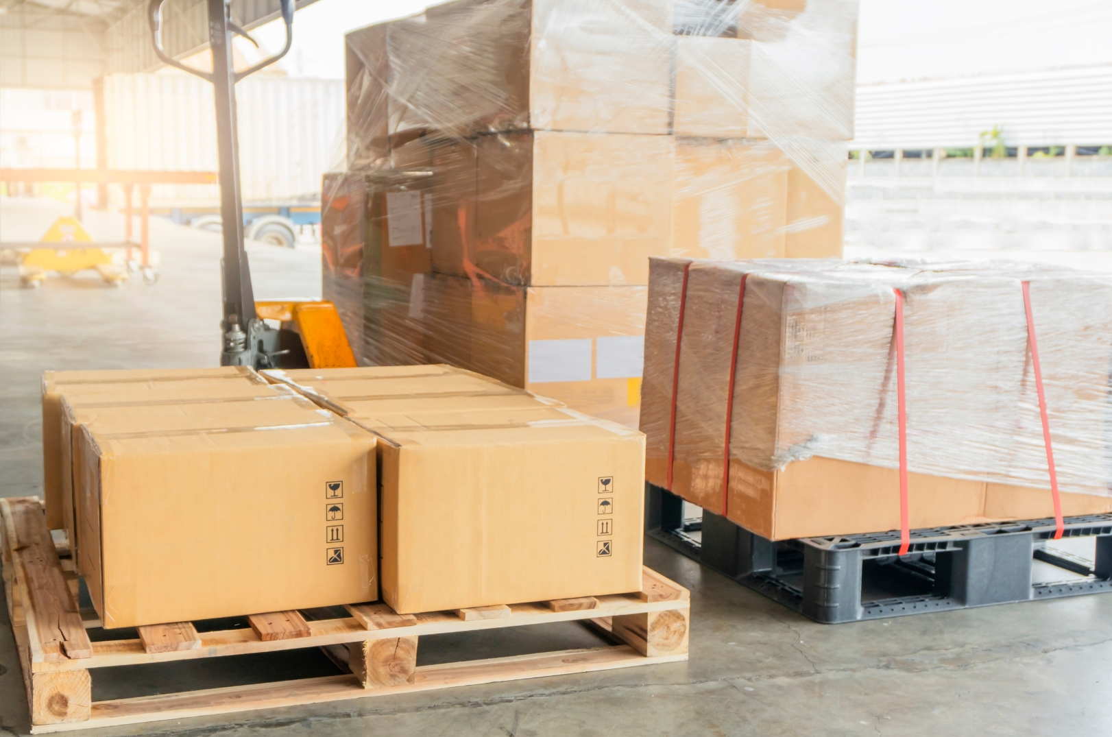 Boxes and pallets in a warehouse, some wrapped in plastic, with a forklift in the background.