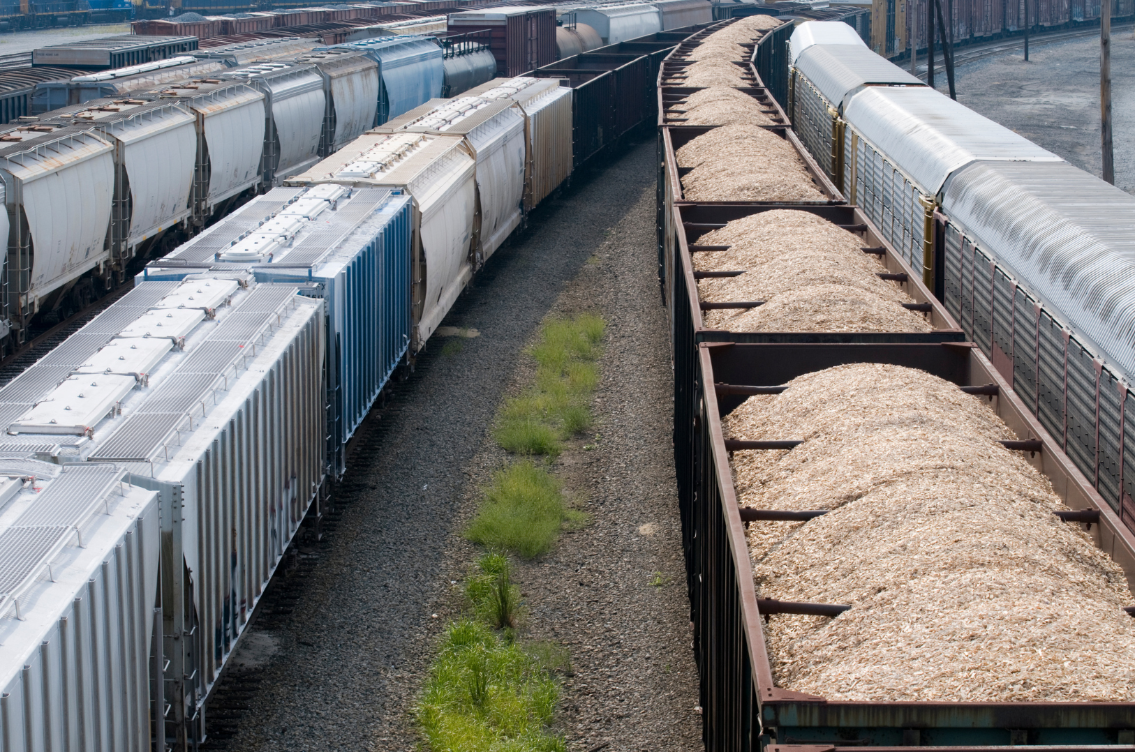 Train cars carrying gravel and other cargo on railroad tracks with small patches of grass in between.