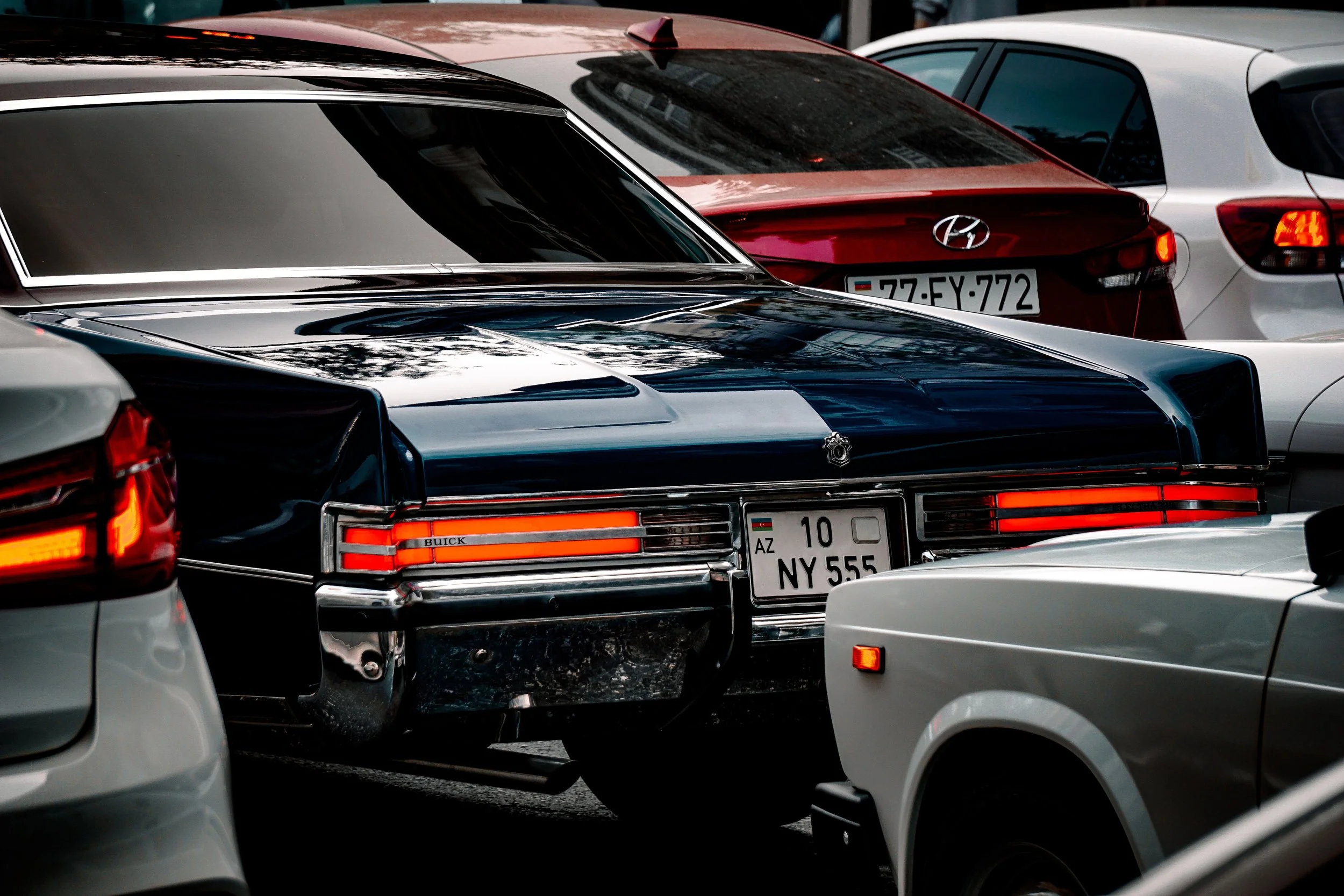 Close-up of several parked cars, including a black vintage Buick and a red Hyundai, with some visible license plates, in a crowded parking lot.