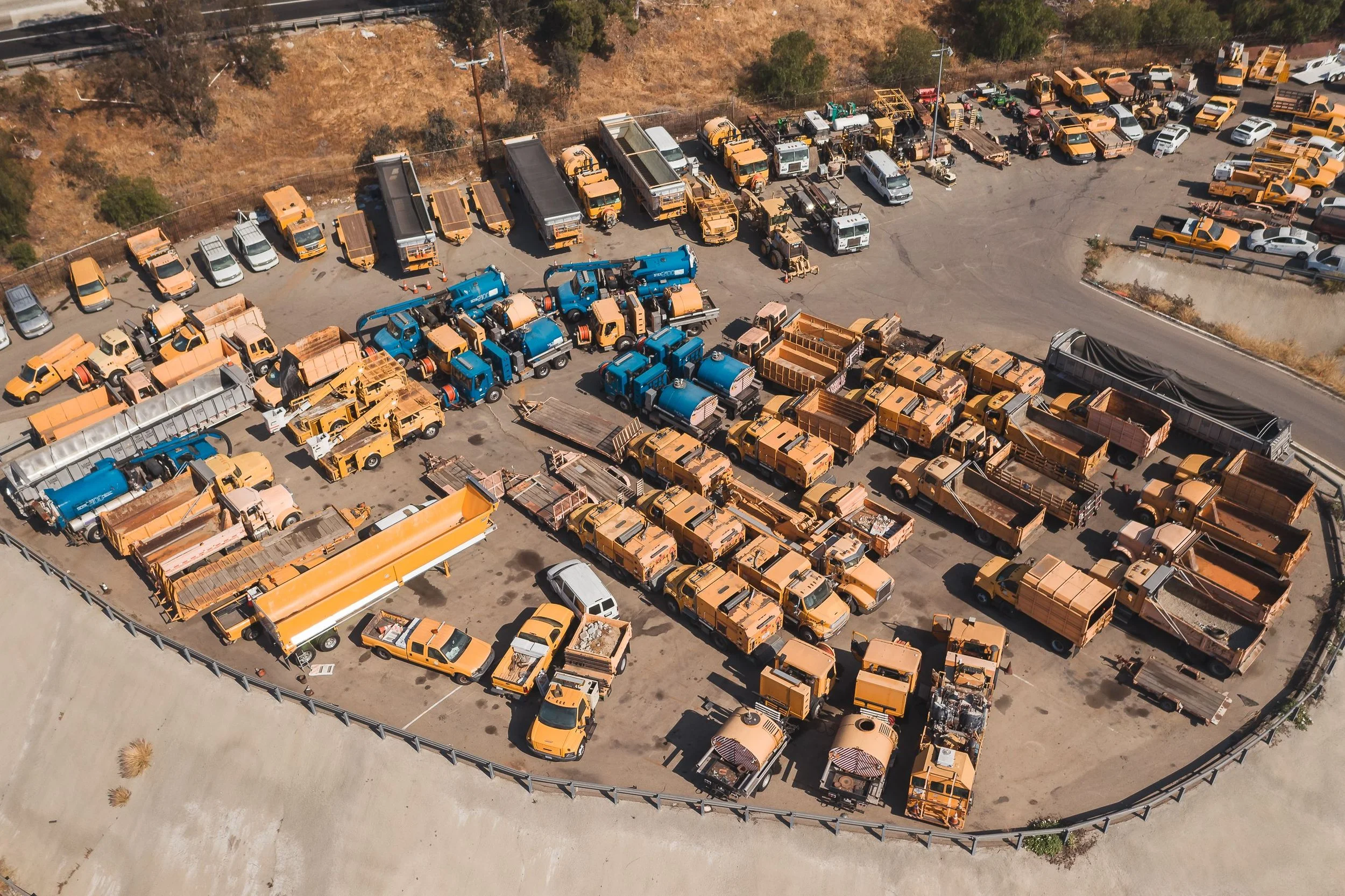 Aerial view of a parking lot filled with various vehicles including utility trucks, service vehicles, and cars, with some vehicles covered or parked near a fenced area. The parking lot is adjacent to a dry, grassy hillside with trees and utility poles.