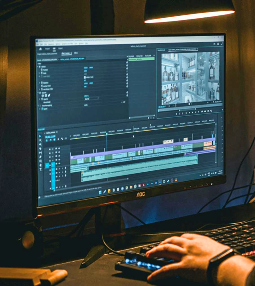 A person working on video editing software on a computer monitor, with a hand operating a control panel, in a dimly lit room.