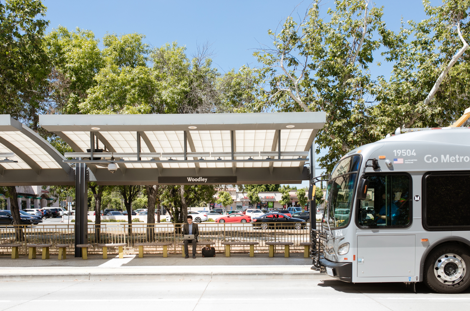Bus stop with a person sitting and using a laptop, several trees, parked cars in the background, and a bus parked at the stop labeled 'Woodley'