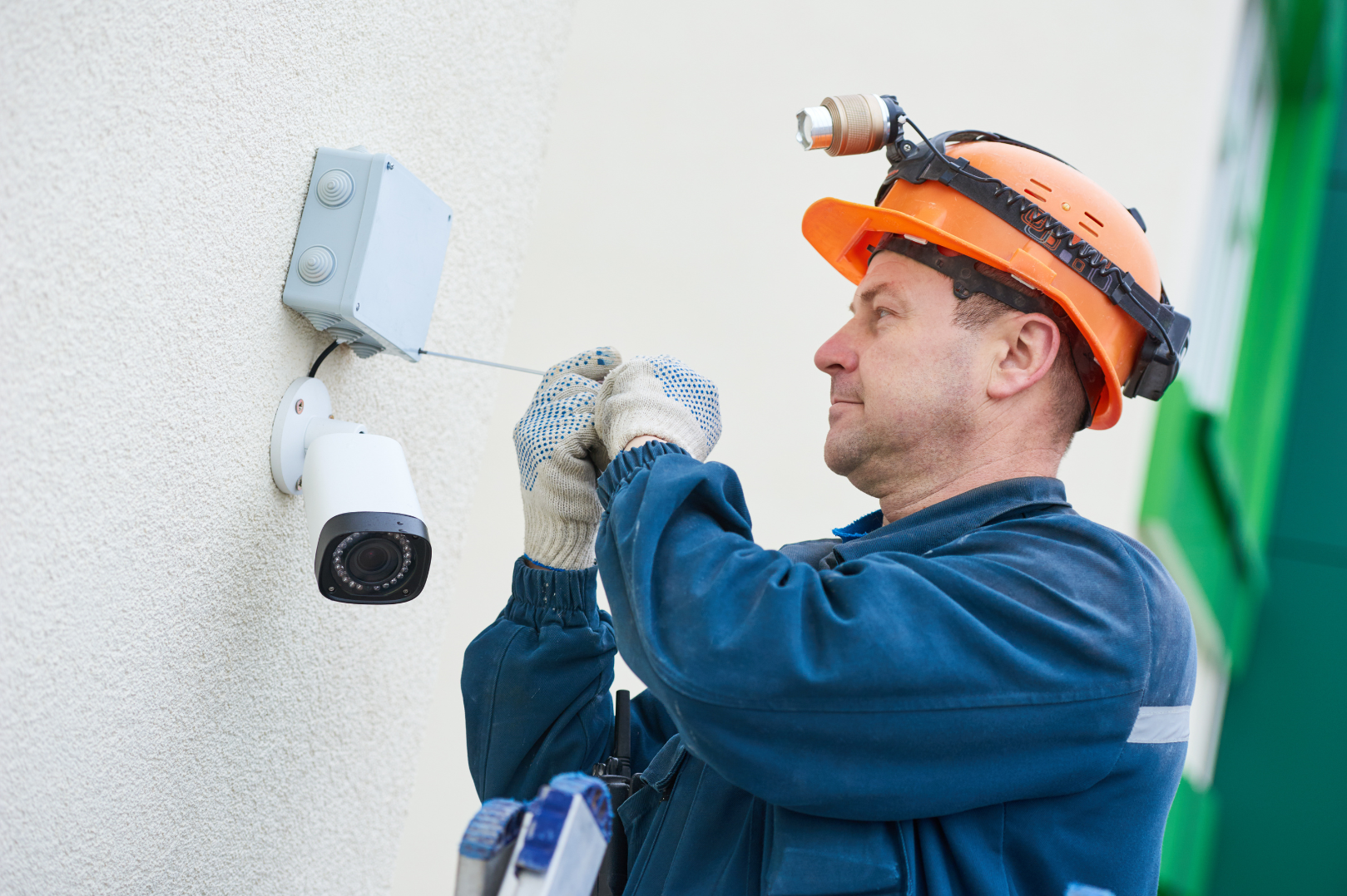 A security camera installer wearing an orange safety helmet with a headlamp and gloves installing a security camera on an exterior wall.