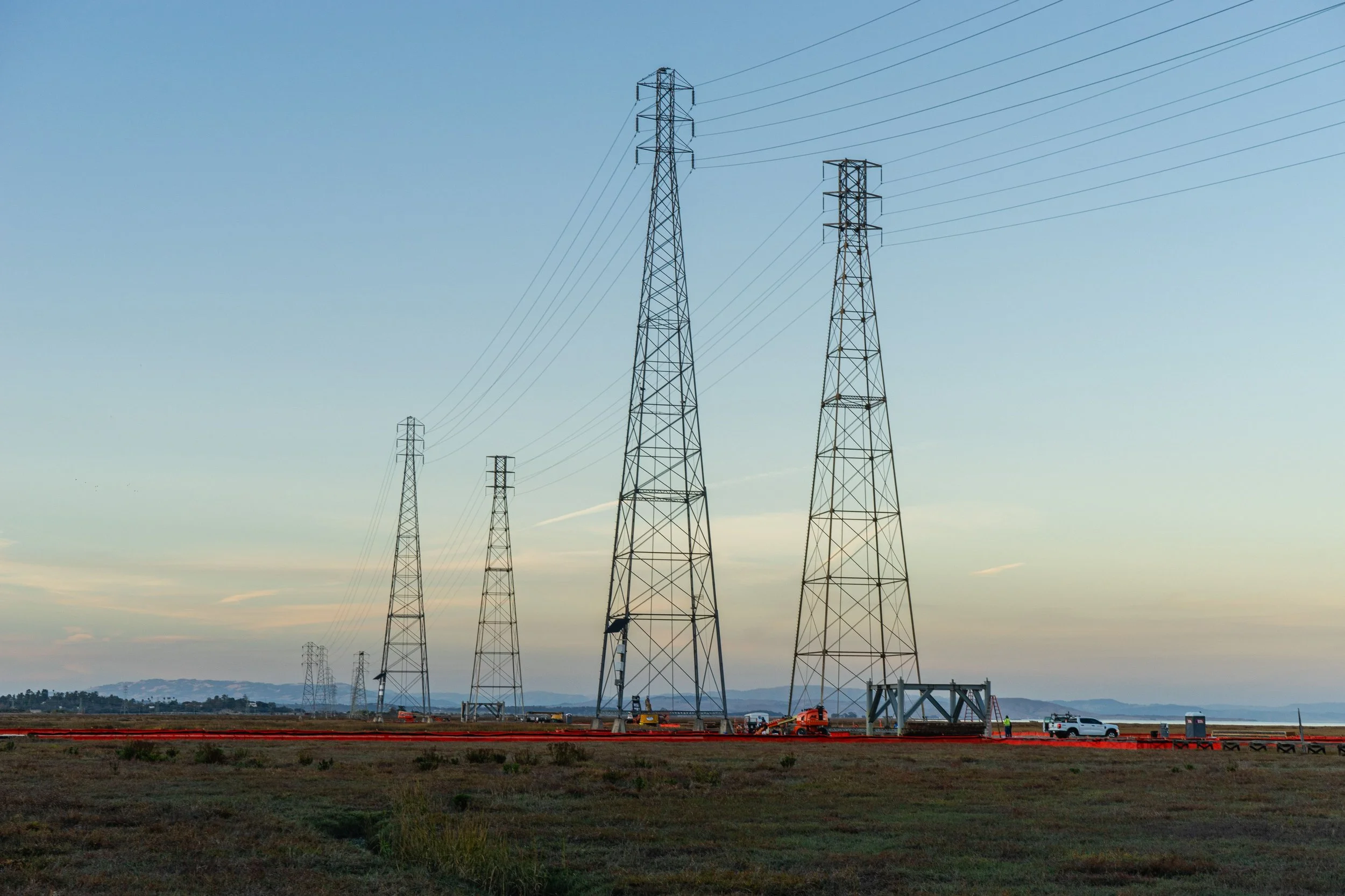 A series of large electrical transmission towers line a landscape at sunset, with a few vehicles and workers visible near the base of the towers.