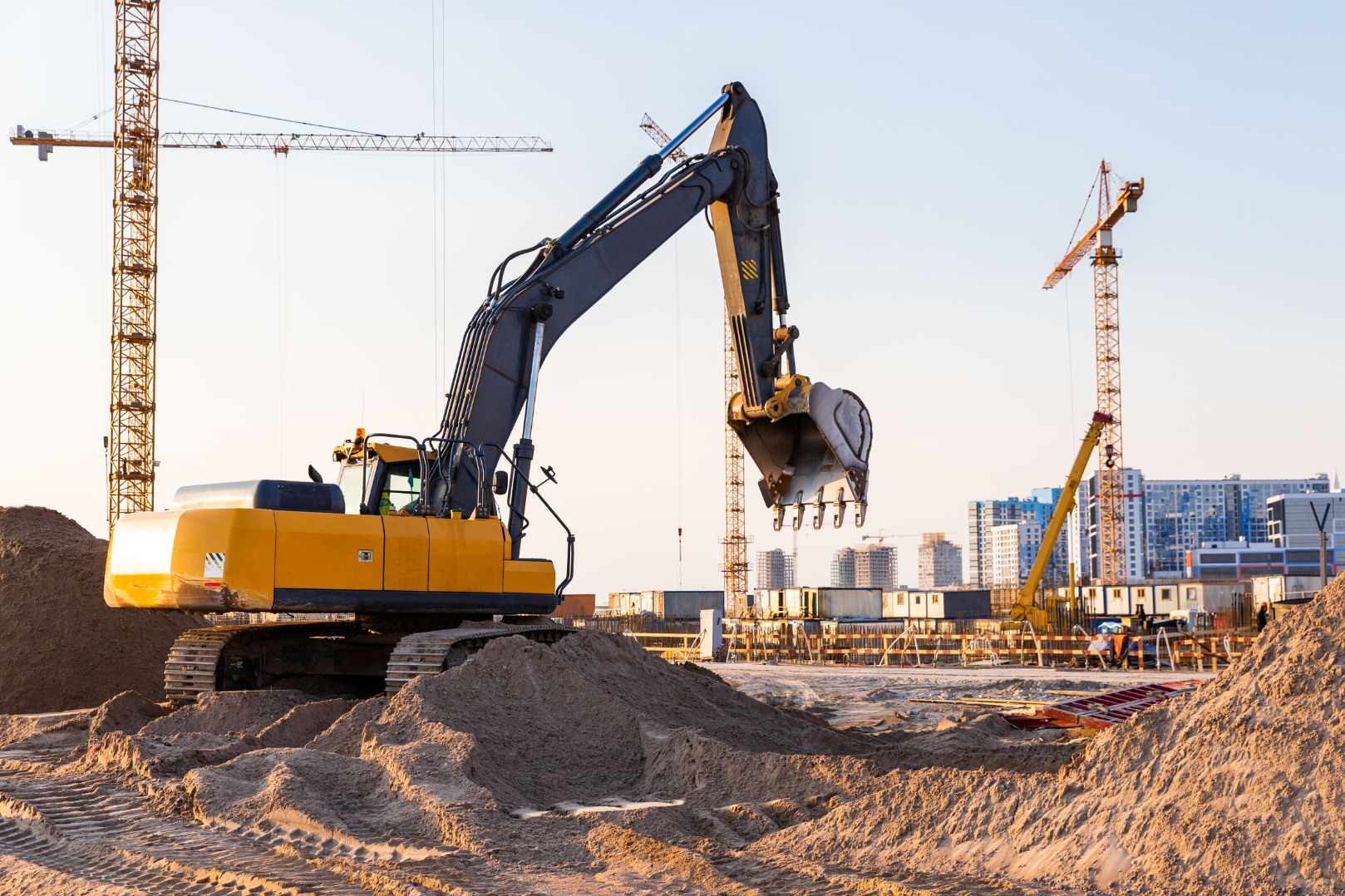 A construction site featuring a yellow excavator with a bucket, surrounded by dirt piles and multiple cranes in the background, with a city skyline in the distance during daylight.